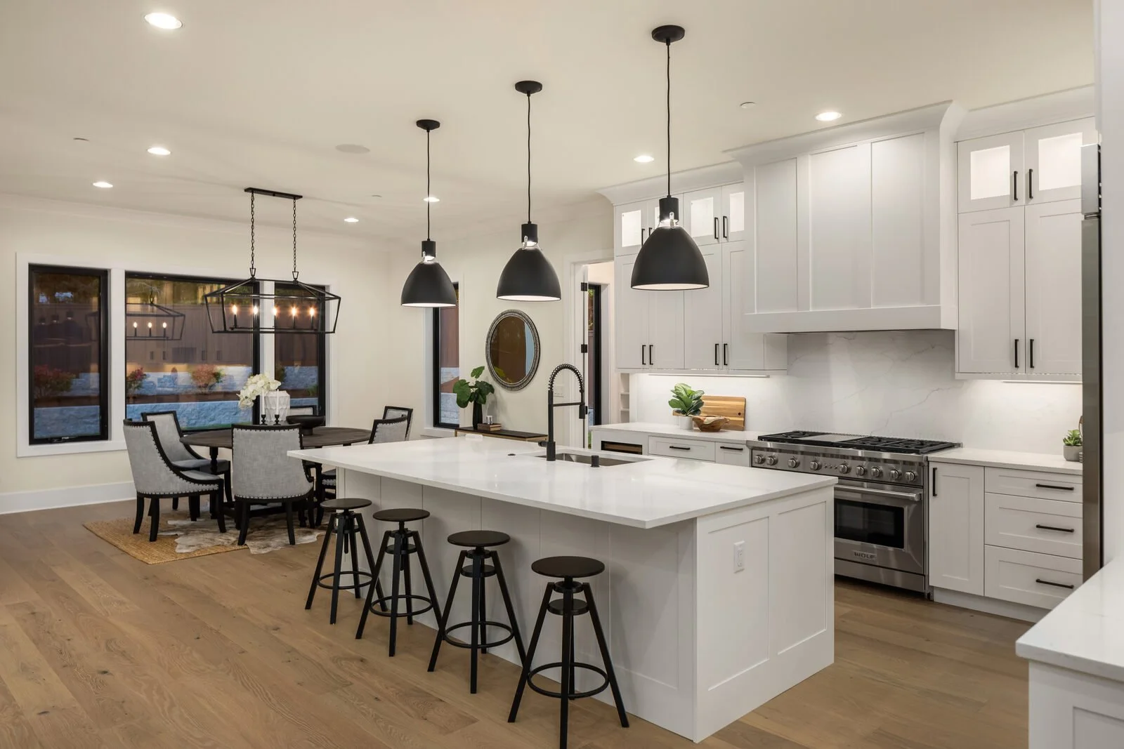 Modern kitchen with white cabinets, a large island with a white countertop, black pendant lights, a stainless steel stove, and a dining area with a table and chairs near windows.