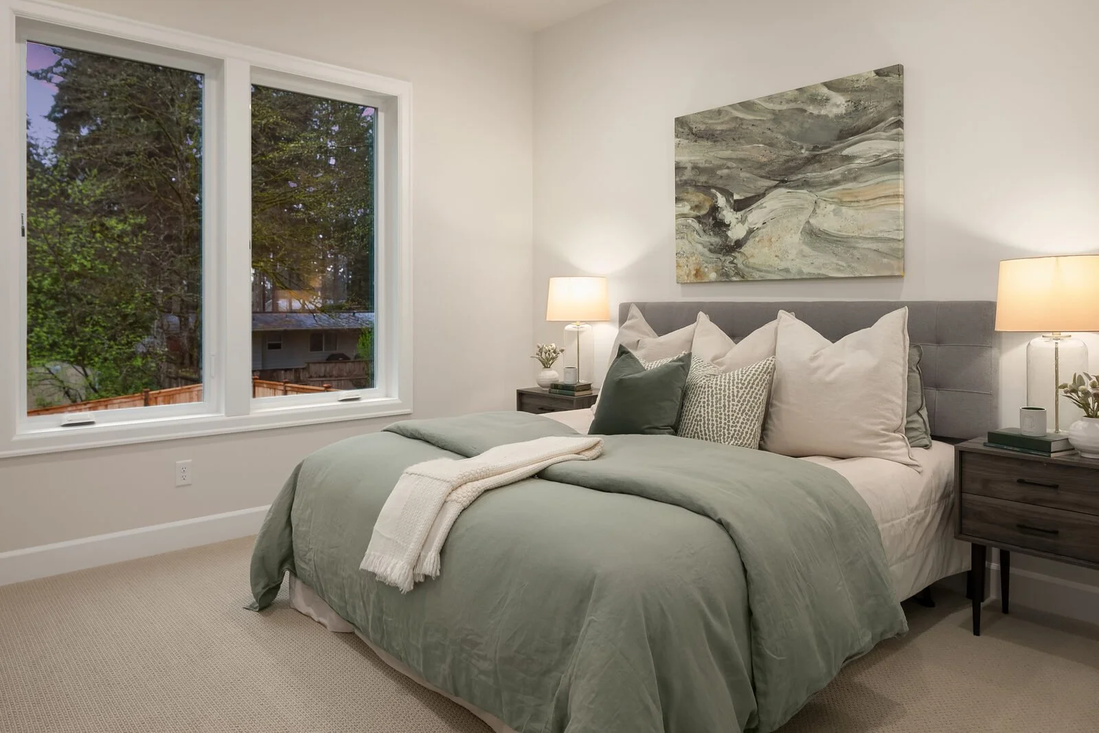 Bedroom with a large window showing trees outside, a bed with green bedding and multiple cushions, two nightstands with lamps, a small plant, and a textured abstract painting above the bed.