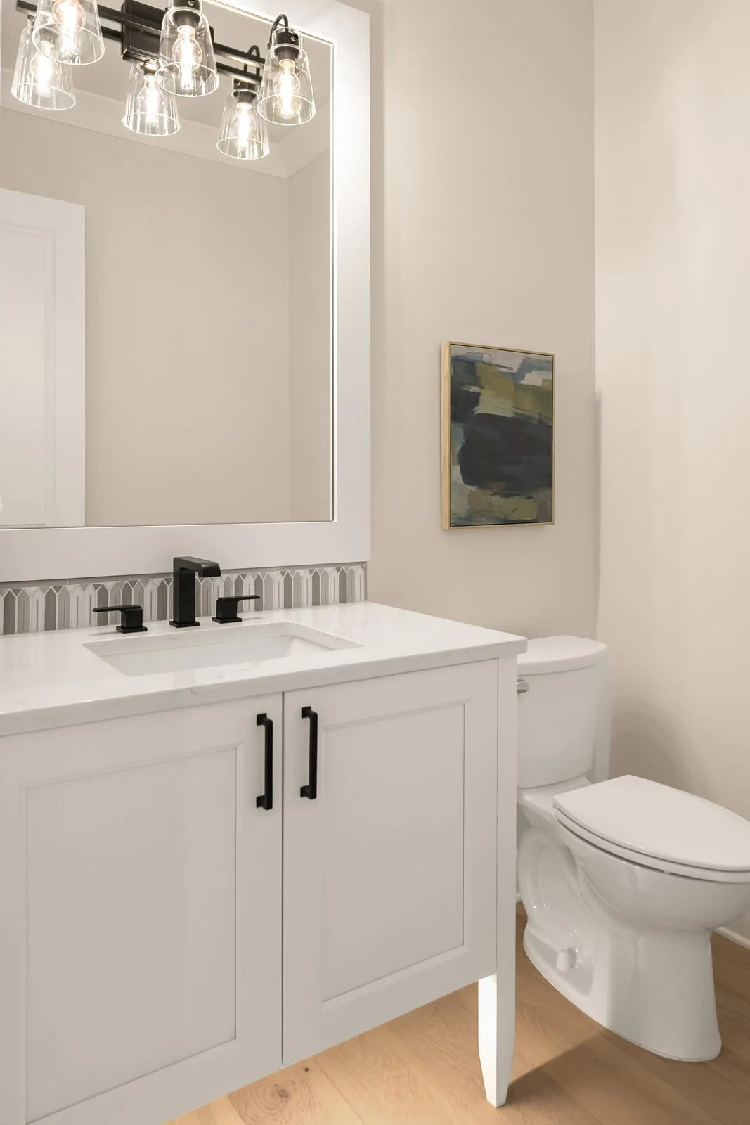 A modern bathroom featuring a white vanity with black handles, a large mirror, black faucet, and a wall-mounted light fixture with multiple glass shades. To the right, there's a toilet and a small framed abstract painting on the wall.
