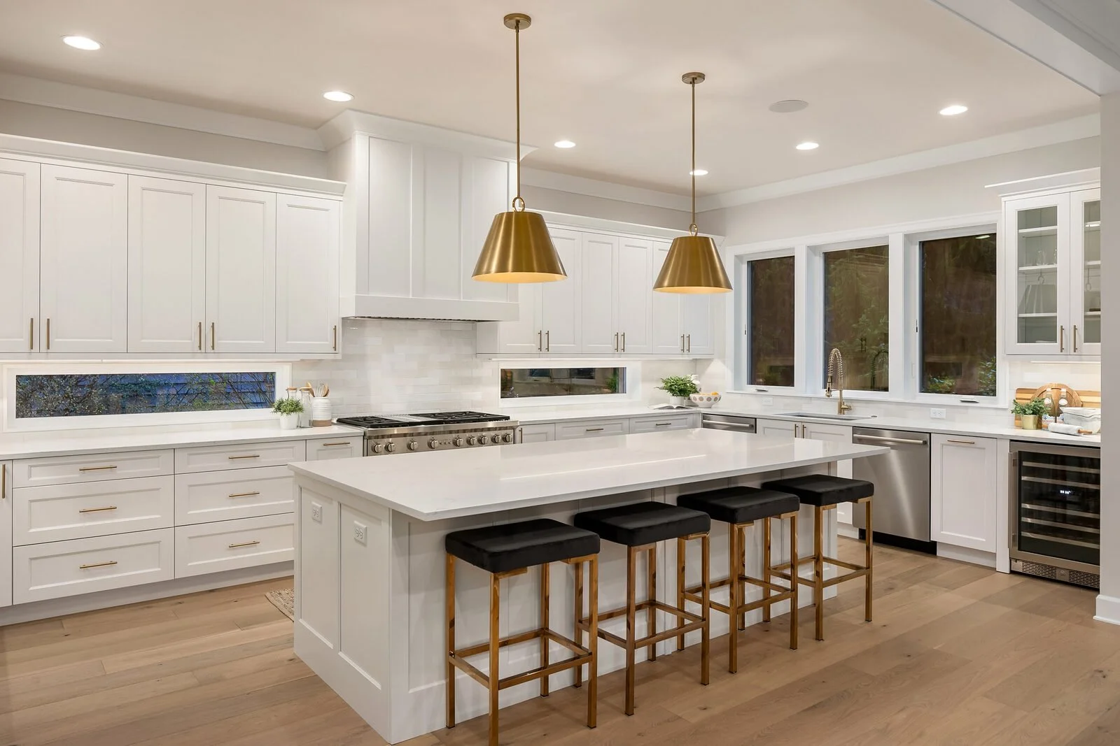 Modern kitchen with white cabinets, a large island with four black and gold stools, and pendant lights. Large windows and hardwood floors.