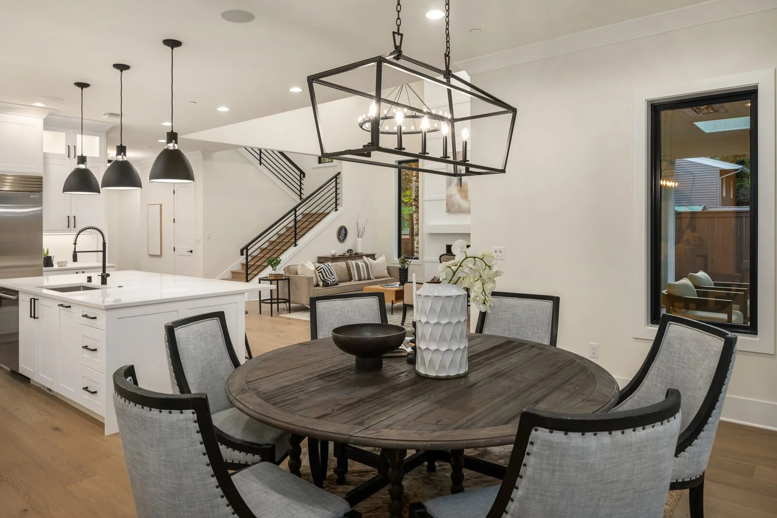 Open-concept living and dining area with a round wooden table, six upholstered chairs, and a decorative chandelier. The space includes a modern kitchen island with three black pendant lights, a beige sofa with cushions, a staircase with black railing