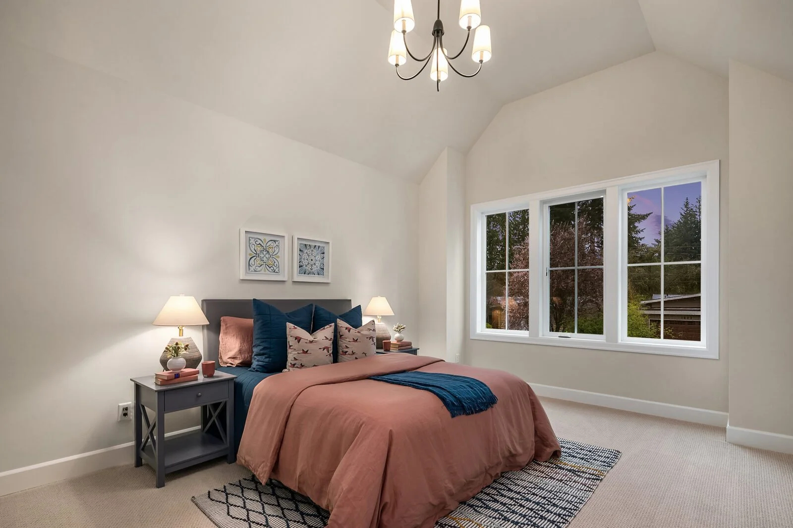 A bedroom with a bed covered in a pinkish-orange duvet, complemented by blue and patterned pillows. There are two nightstands with lamps on each side of the bed, and large windows showing a view of trees outside. White walls and a ceiling with a mode
