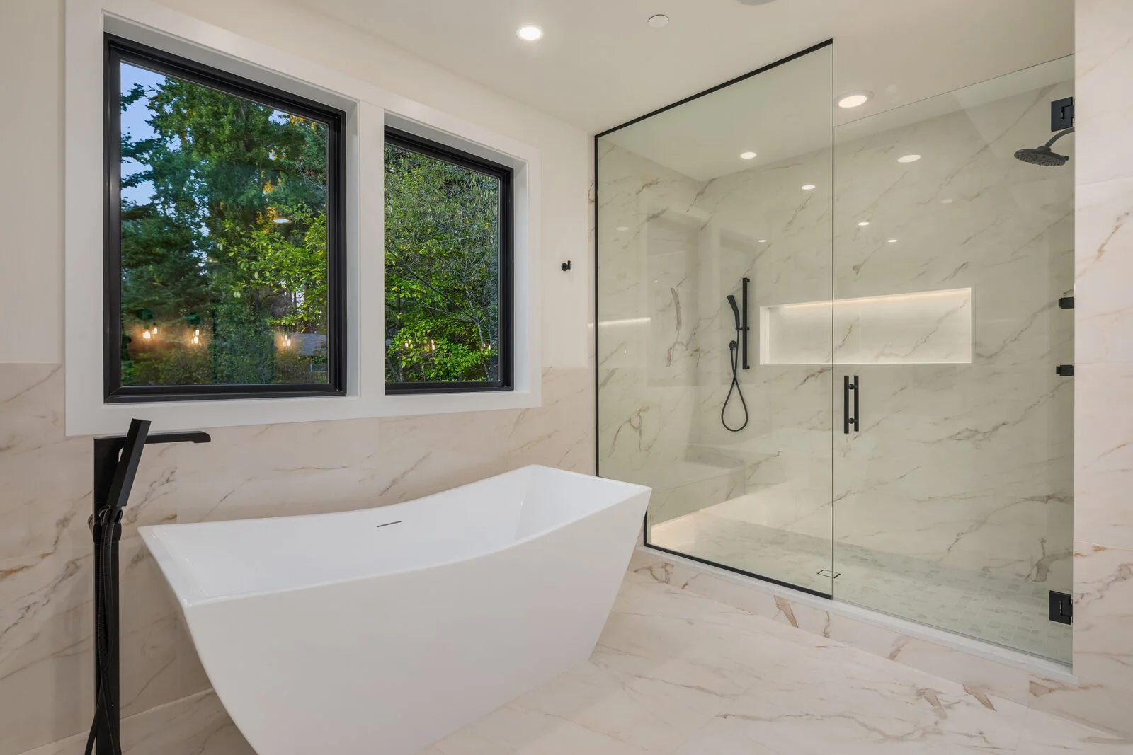 Modern bathroom with a freestanding bathtub, large windows showing greenery outside, and a glass-enclosed walk-in shower with marble walls and black fixtures.