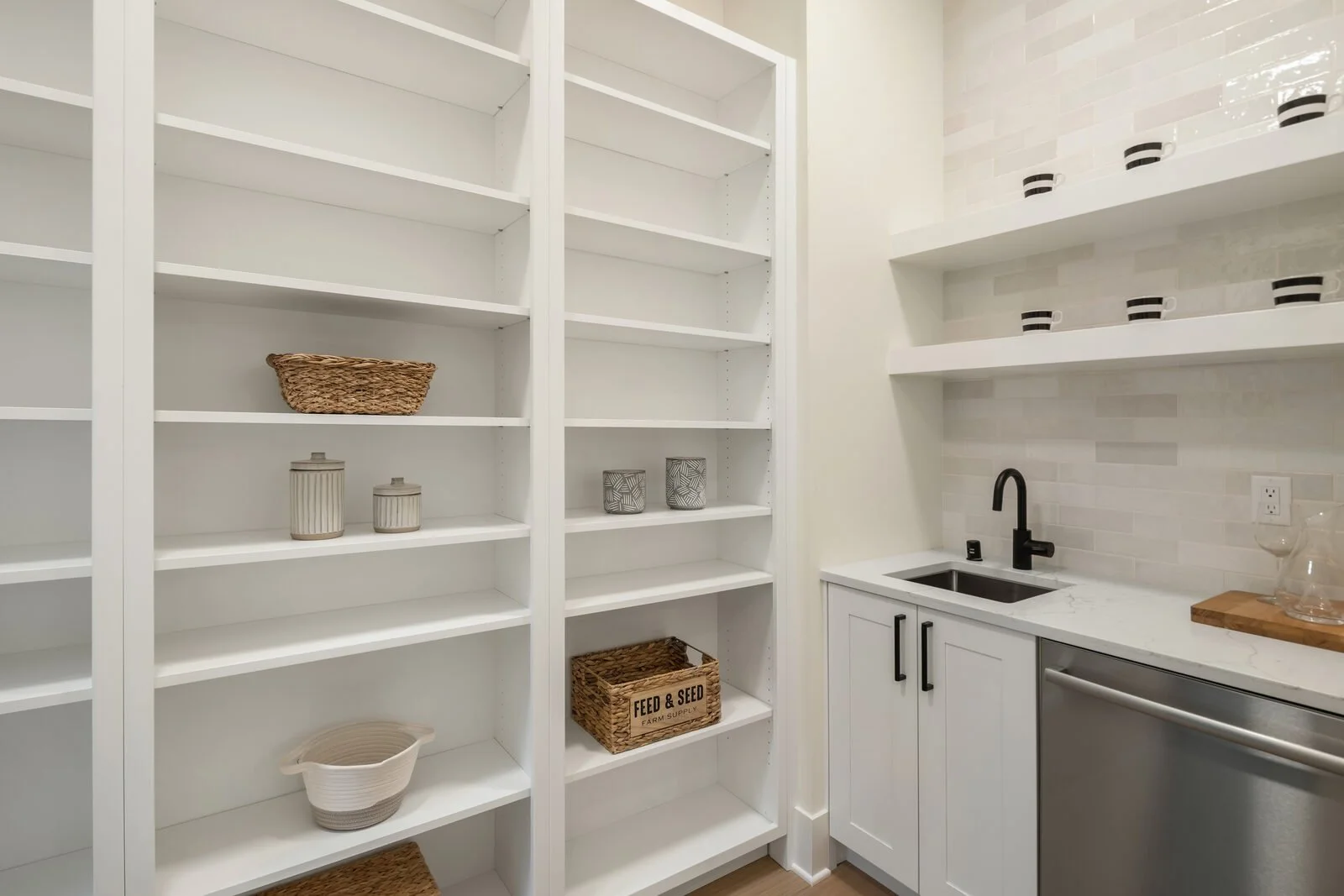 Empty white kitchen shelves with baskets and jars, small sink with black faucet, marble countertop, and open shelving with cups.