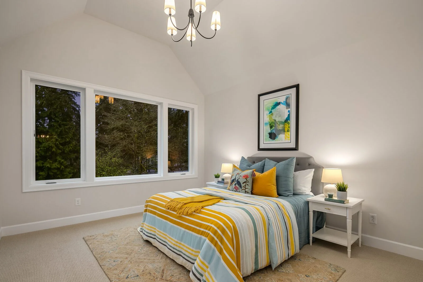 A bright bedroom with white walls and a large window showing trees outside. There is a bed with a colorful striped bedding set, decorative pillows including yellow, blue, and a pillow with a floral pattern. Two white nightstands with lamps and small 