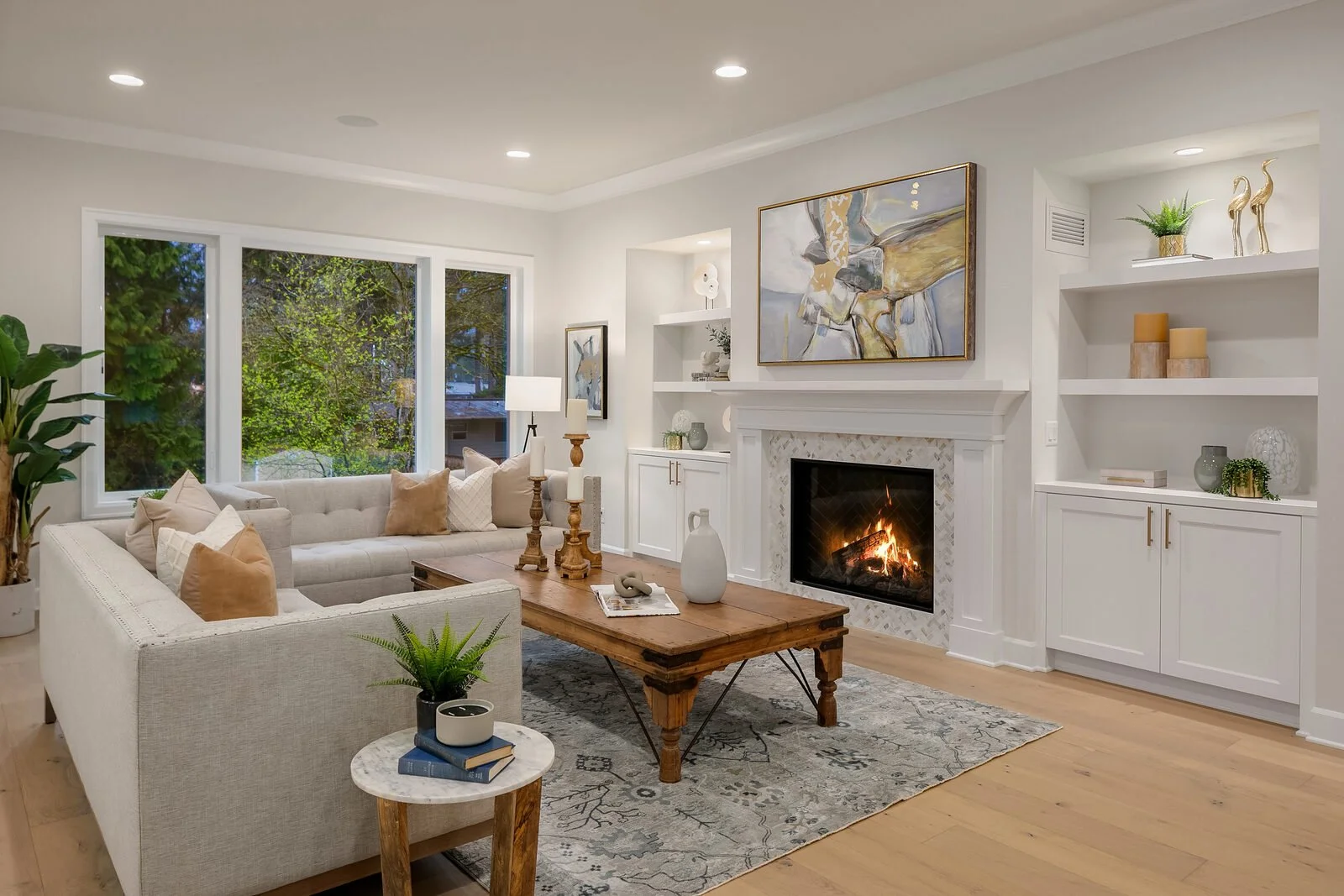 Living room with large window, white couch with beige and white pillows, wooden coffee table with candles, white fireplace with abstract painting, built-in white shelves with decor, houseplant, and natural light.