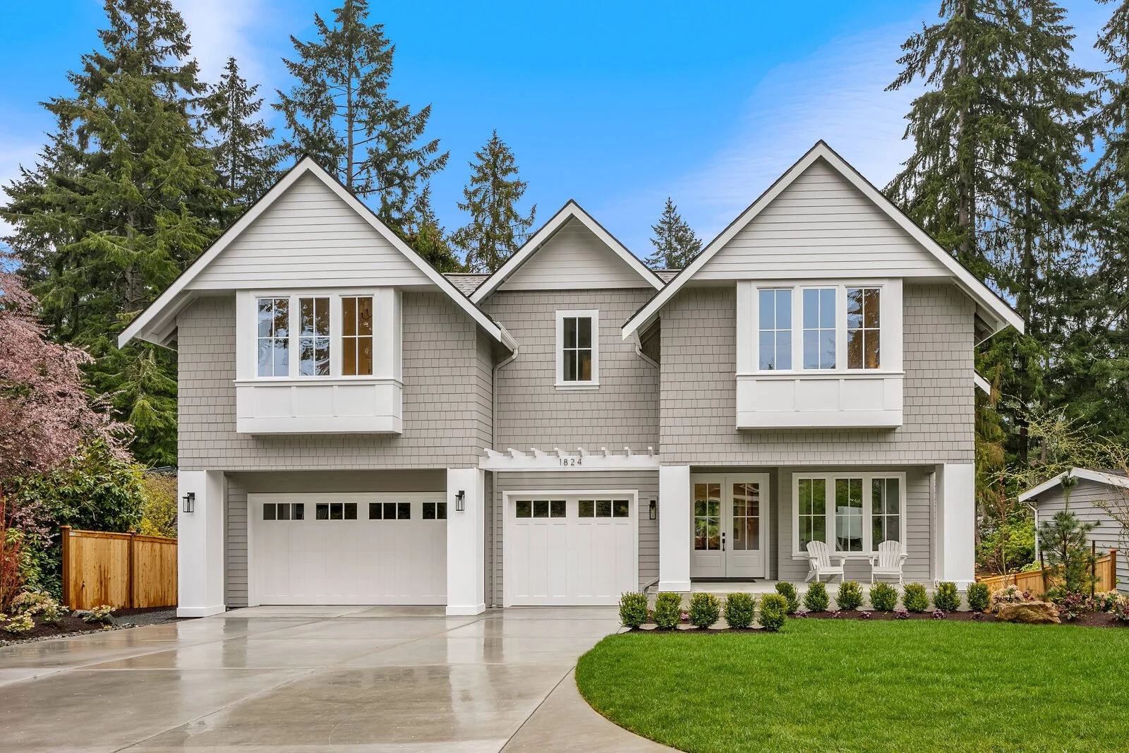 A modern two-story house with gray siding, white trim, and a gable roof, surrounded by greenery and trees, with a manicured lawn and a concrete driveway in front.