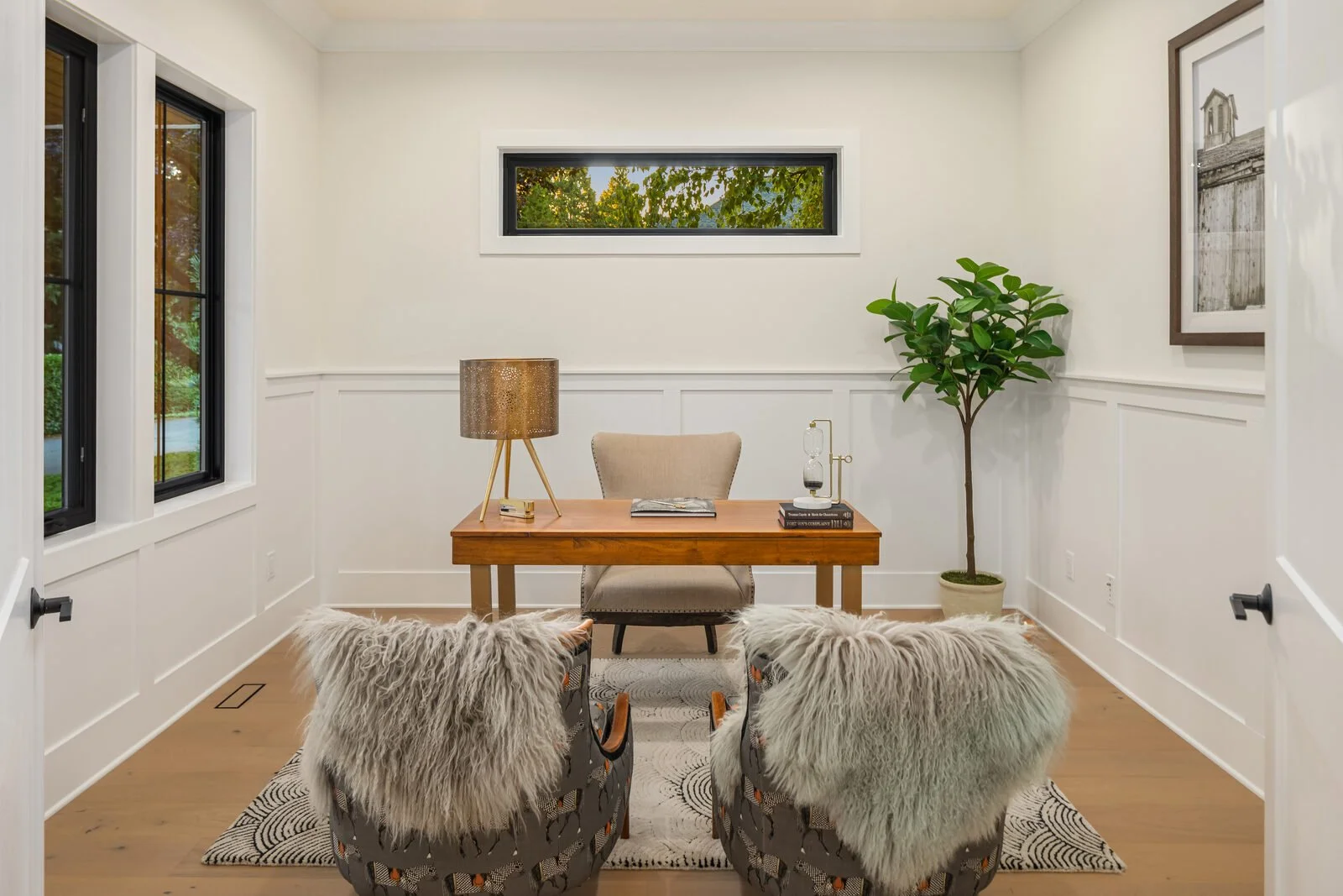 A home office with a wooden desk, two patterned chairs with fur throws, a beige upholstered chair behind the desk, a table lamp, a book, a small glass lamp, a tall potted plant, and framed wall art. There is a window showing greenery outside and the 