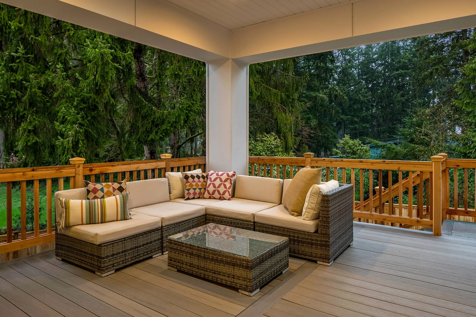 Outdoor covered porch with sectional wicker sofa, colorful patterned pillows, glass-top coffee table, wooden railing, and lush green trees in the background.