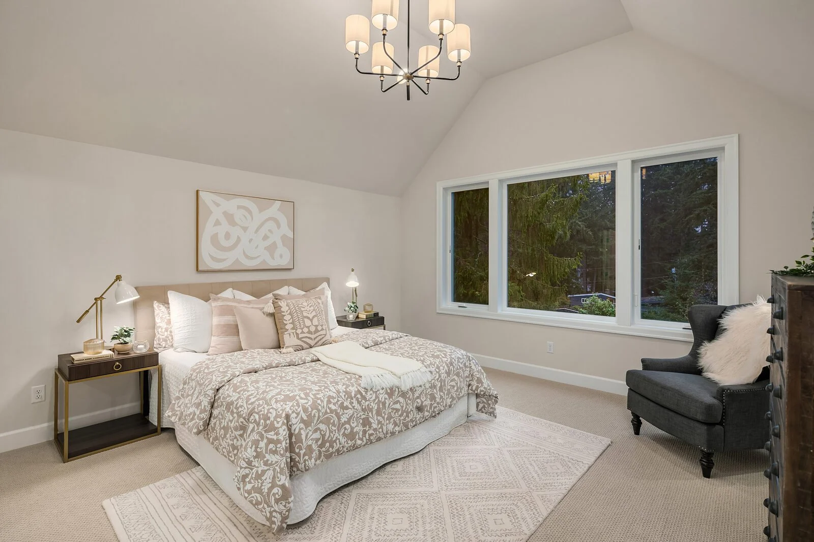 A cozy bedroom with a large bed featuring light patterned bedding, several pillows, and a cream-colored throw. Two modern bedside tables with lamps flank the bed. The room has a large window showing trees outside, a plush dark gray armchair with a wh