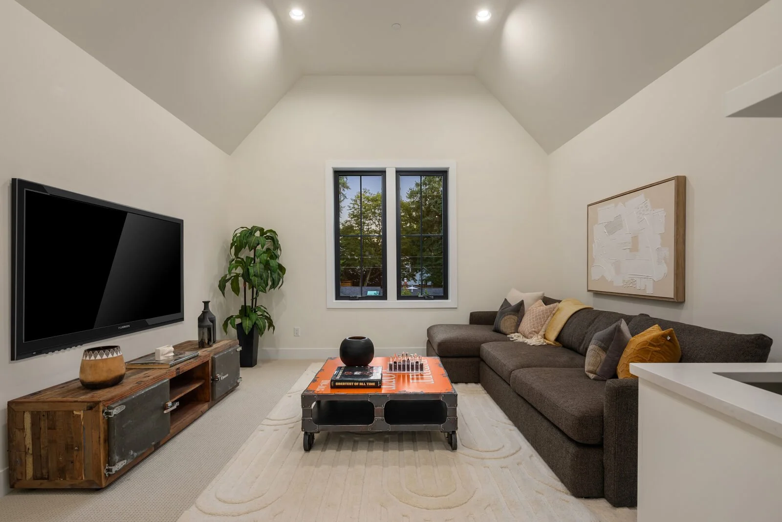 Living room with a dark sectional sofa, window with trees outside, TV on a wooden stand, potted plant, abstract artwork, coffee table with books and decor, and pillows.