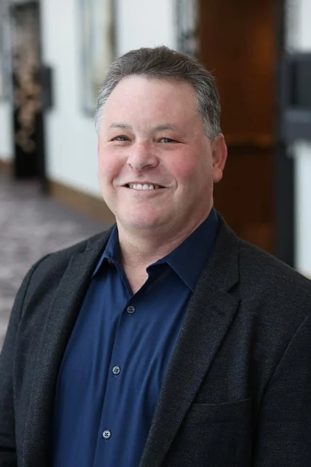 A smiling man in a dark blazer and blue collared shirt, standing indoors with a blurred background.