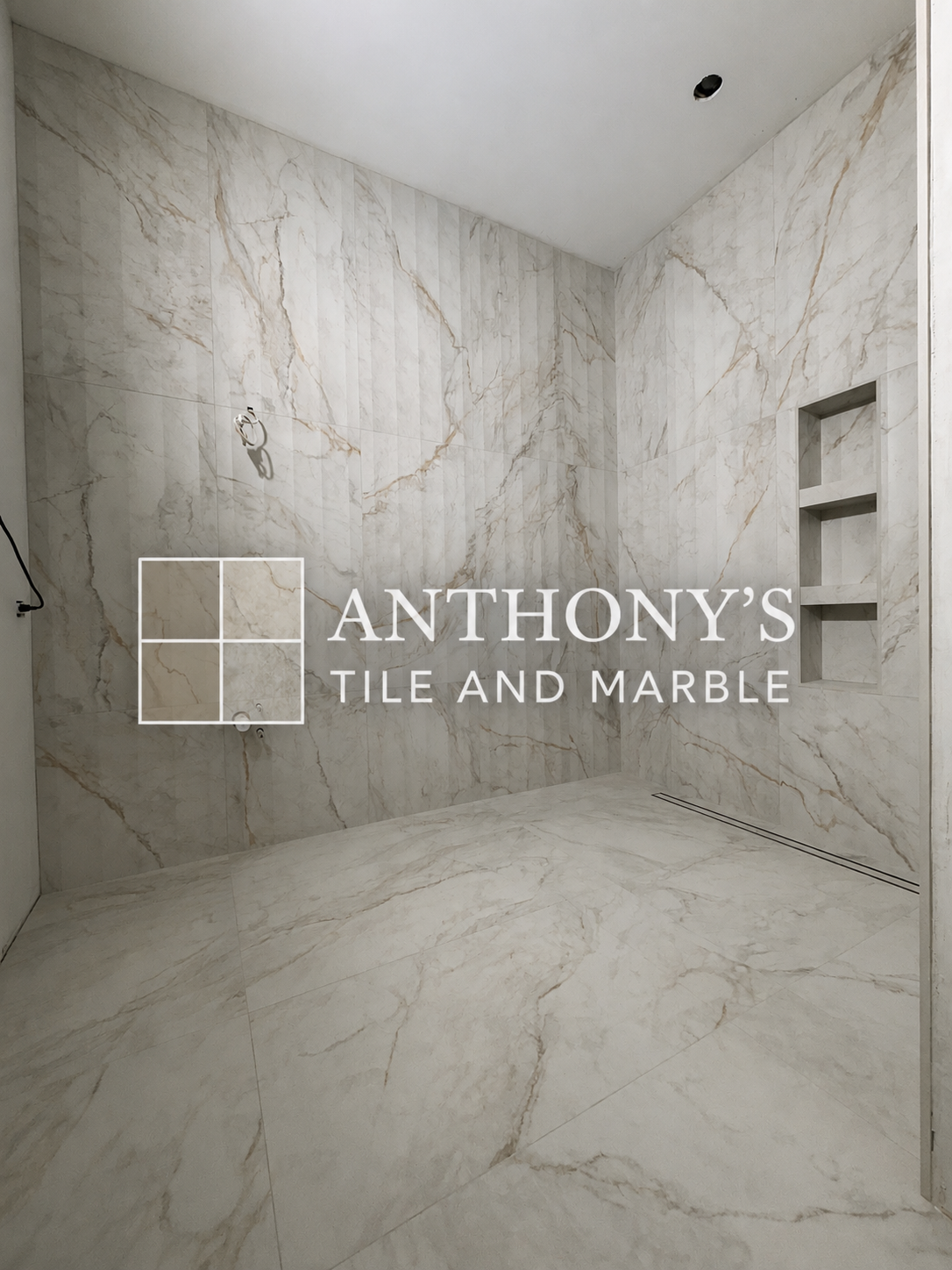 A bathroom with marble tile walls and floor, built-in wall shelves, and a ceiling outlet for a light fixture. The image has the logo of Anthony’s Tile and Marble.