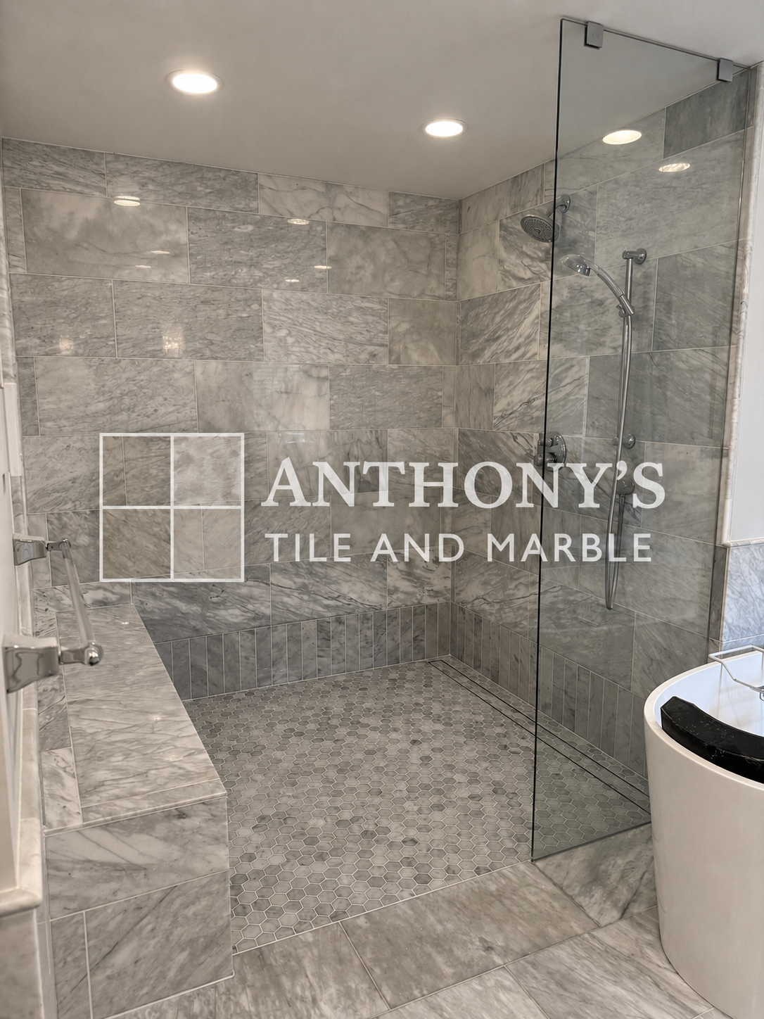 Showers with gray marble tiles and a glass divider, with a toilet in the foreground, in a bathroom with white ceiling and recessed lighting, with the logo of Anthony's Tile and Marble overlaid.