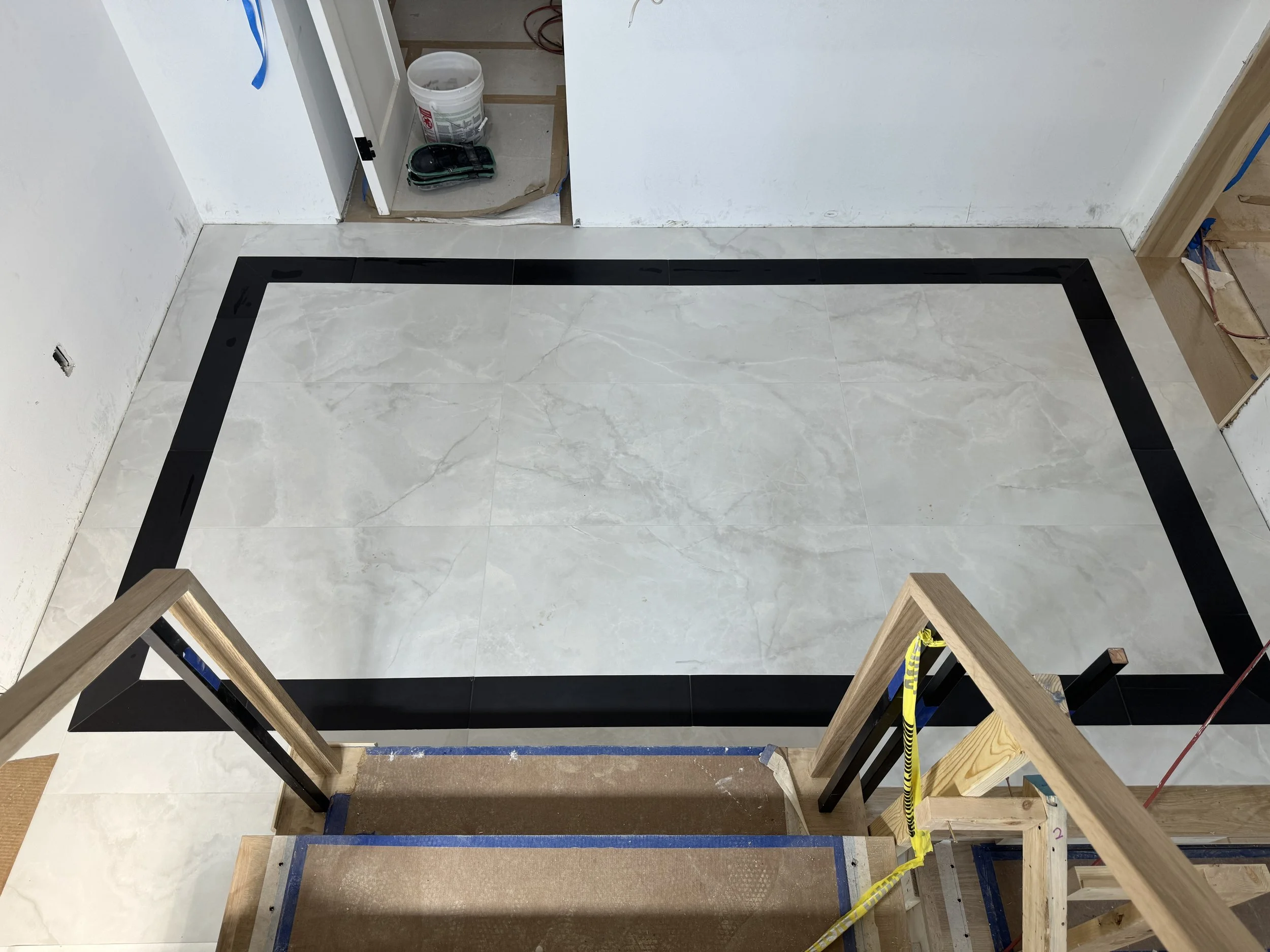 Under construction room with newly installed light-colored marble tile flooring, bordered with black tiles, and wooden framing at the entrance with construction tools and supplies visible in the background.
