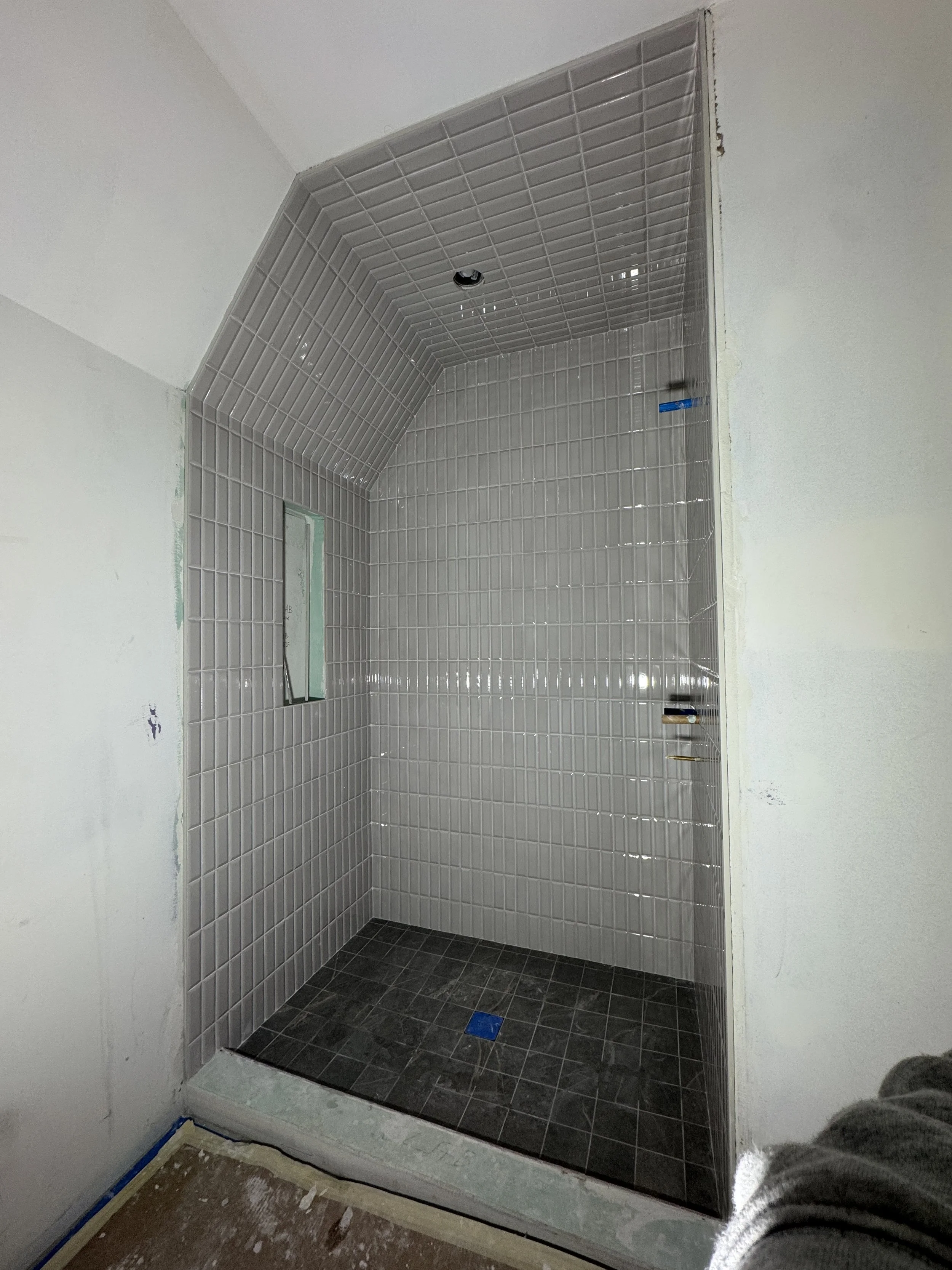 A tiled shower stall under construction with gray tiles on the walls and black tiles on the floor. There is a small tile niche and an opening in the wall, with visible plumbing.