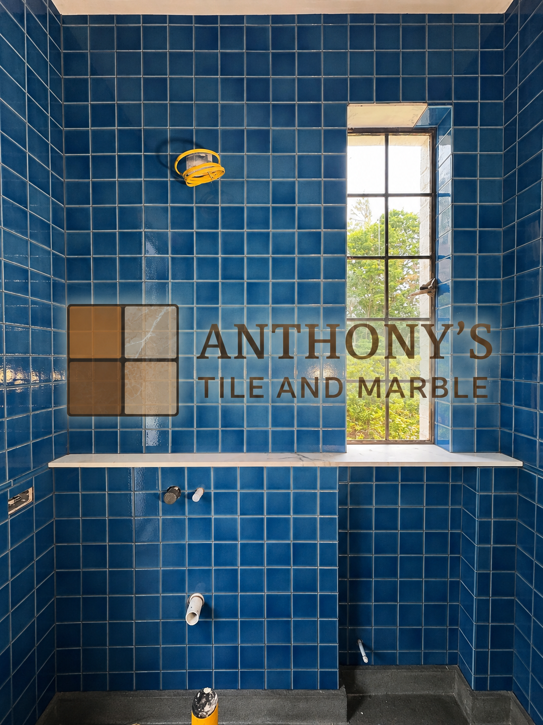 Interior of a bathroom under renovation with blue tiled walls, a window showing greenery outside, and plumbing fixtures not yet installed.