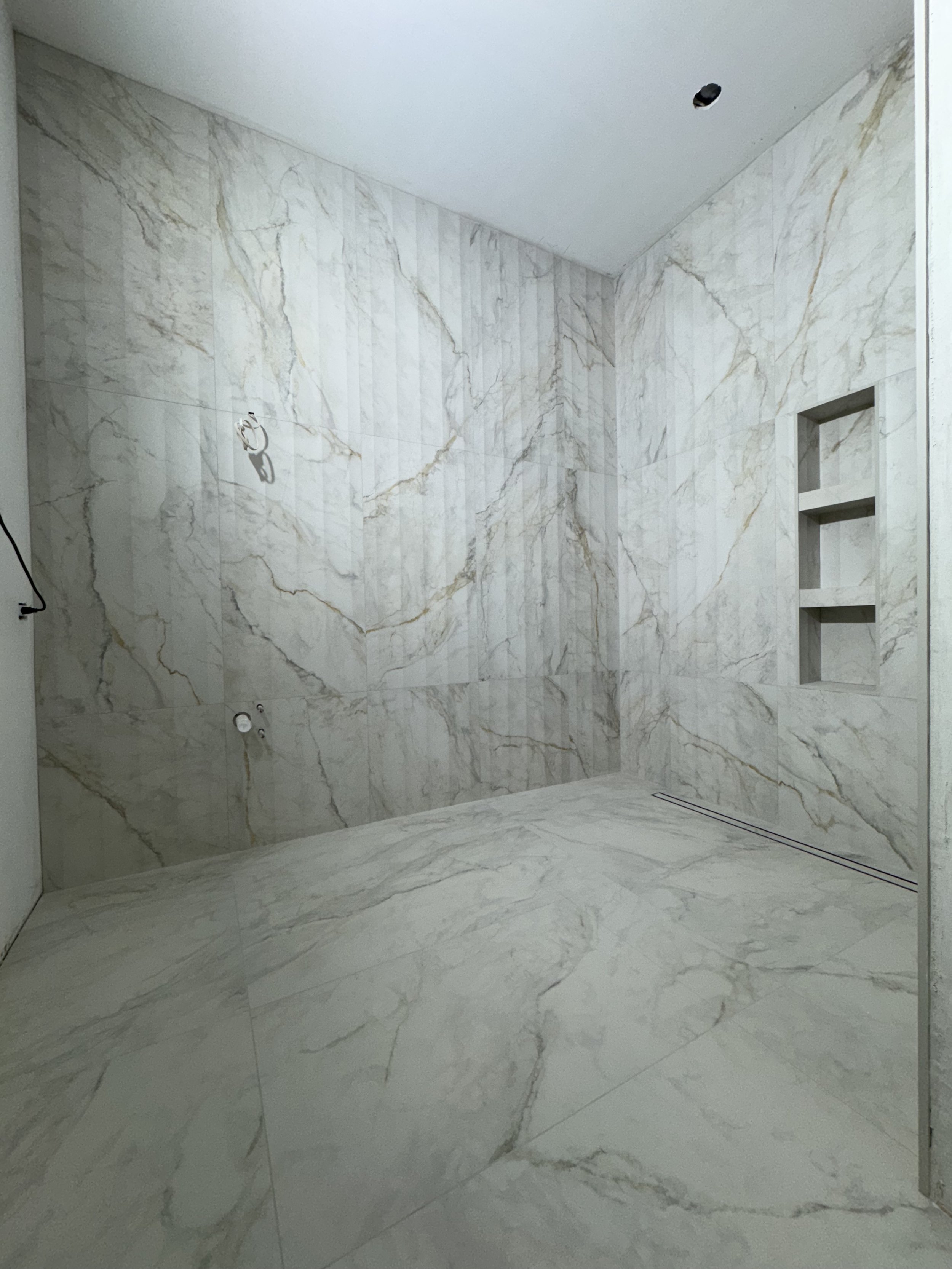 Empty walk-in shower with marble tiles on walls and floor, built-in shelves on the right wall, and a ceiling vent.