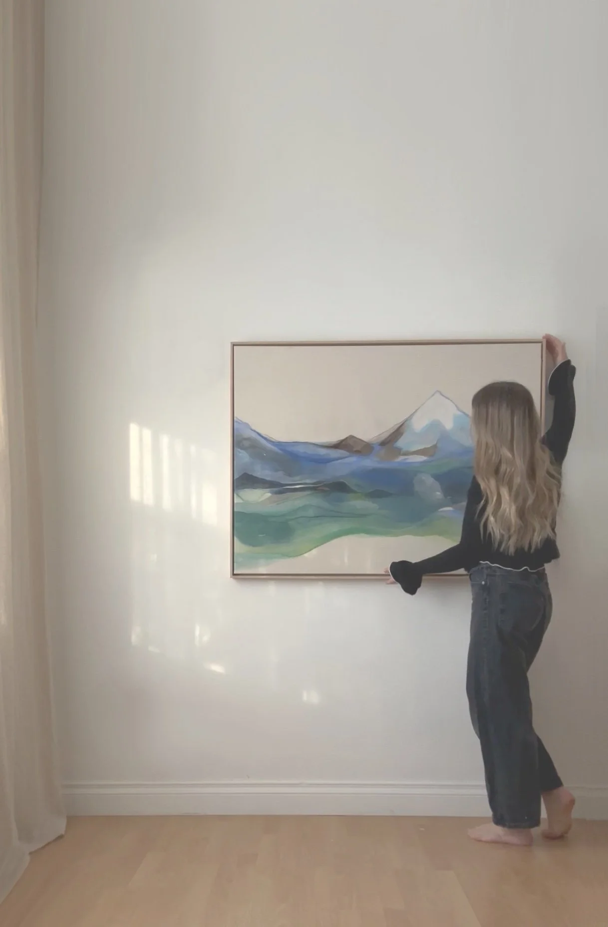 Person hanging a framed watercolor painting of mountains and lakes on a plain white wall.