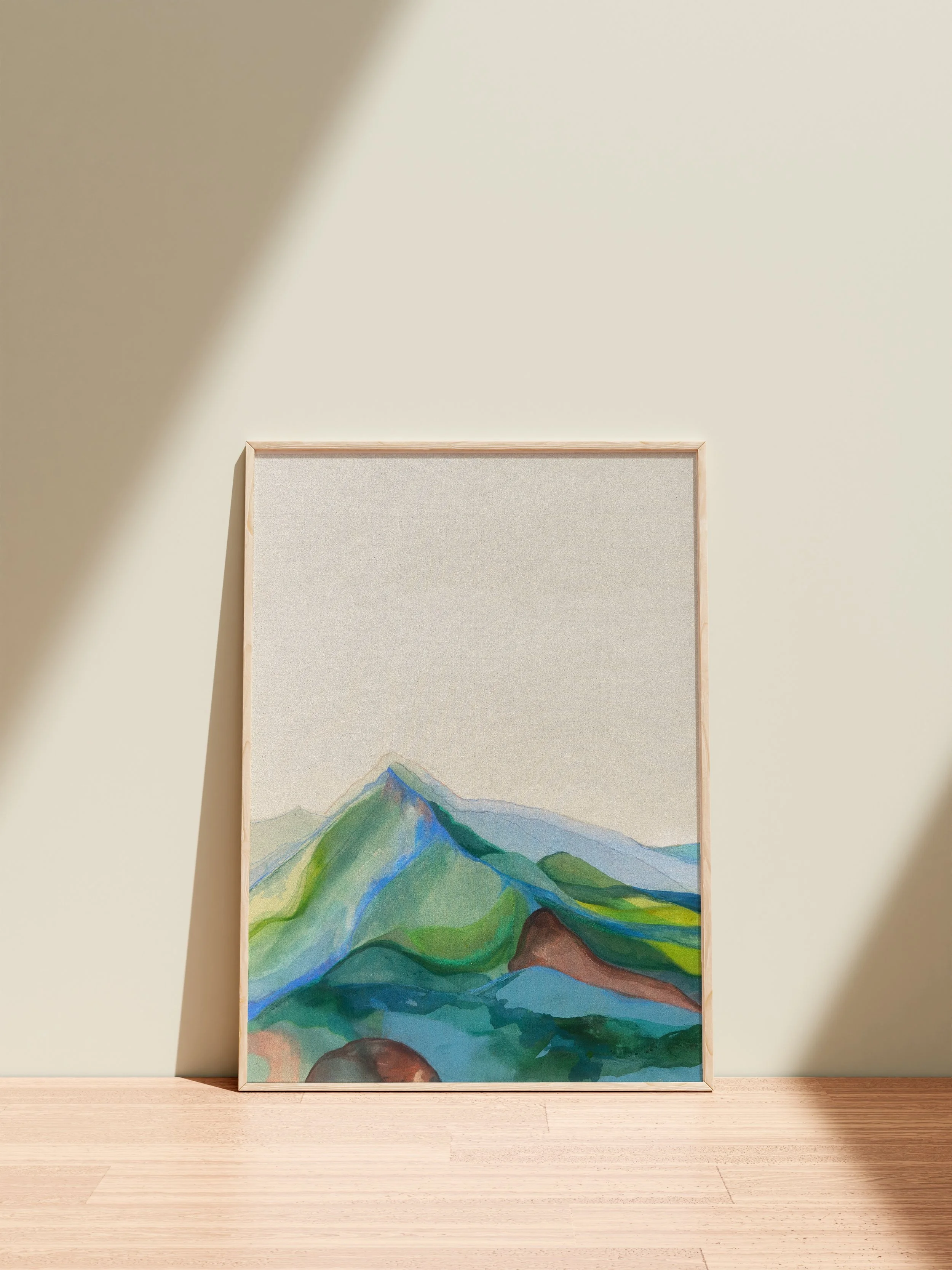 A framed watercolor landscape painting of mountains with green and blue hues, leaning against a light-colored wall on a wooden floor, with sunlight casting shadows on the wall.