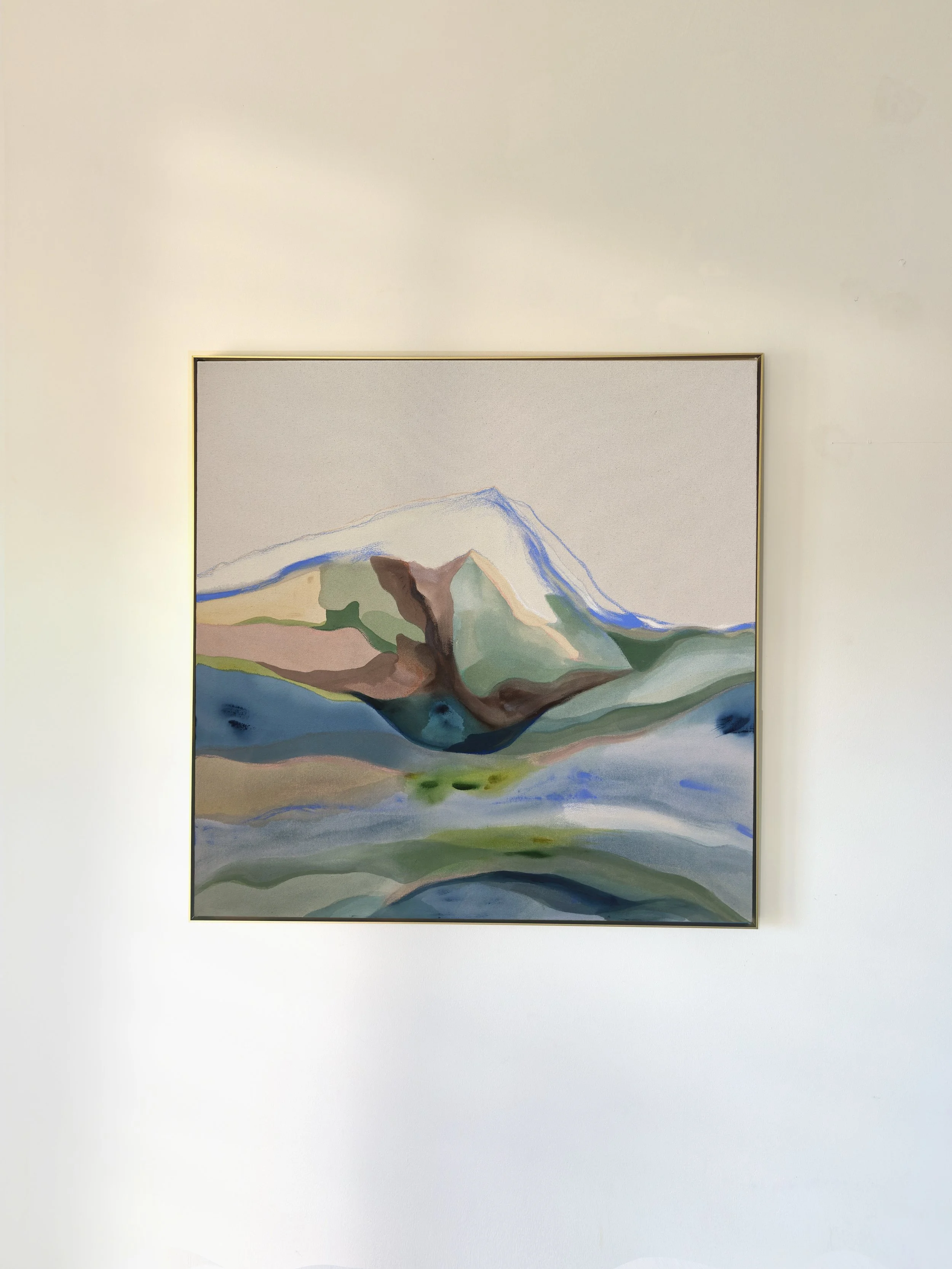 An abstract landscape painting of mountains and water in pastel and earth tones, framed and hanging on a beige wall.