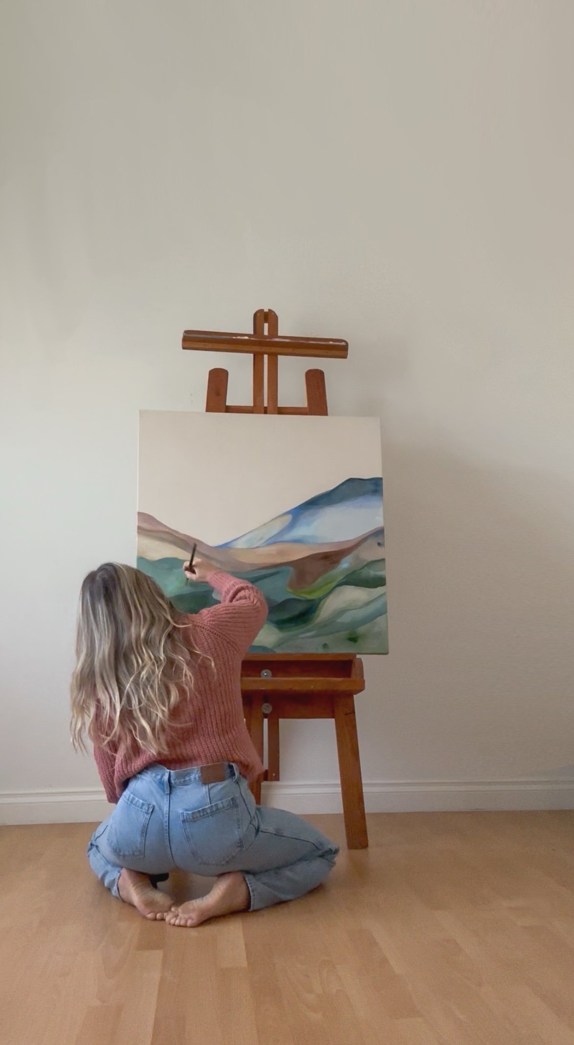 A woman kneeling on the floor, painting an abstract landscape on a canvas mounted on an easel. The painting features mountains and valleys in various shades of blue, green, and brown.