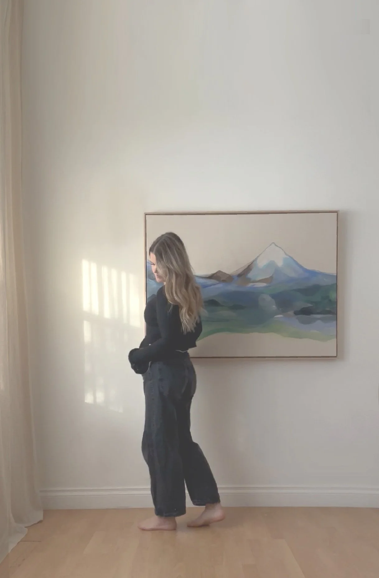 A woman with long hair standing barefoot near a light-colored wall with a framed painting of a mountain landscape behind her.