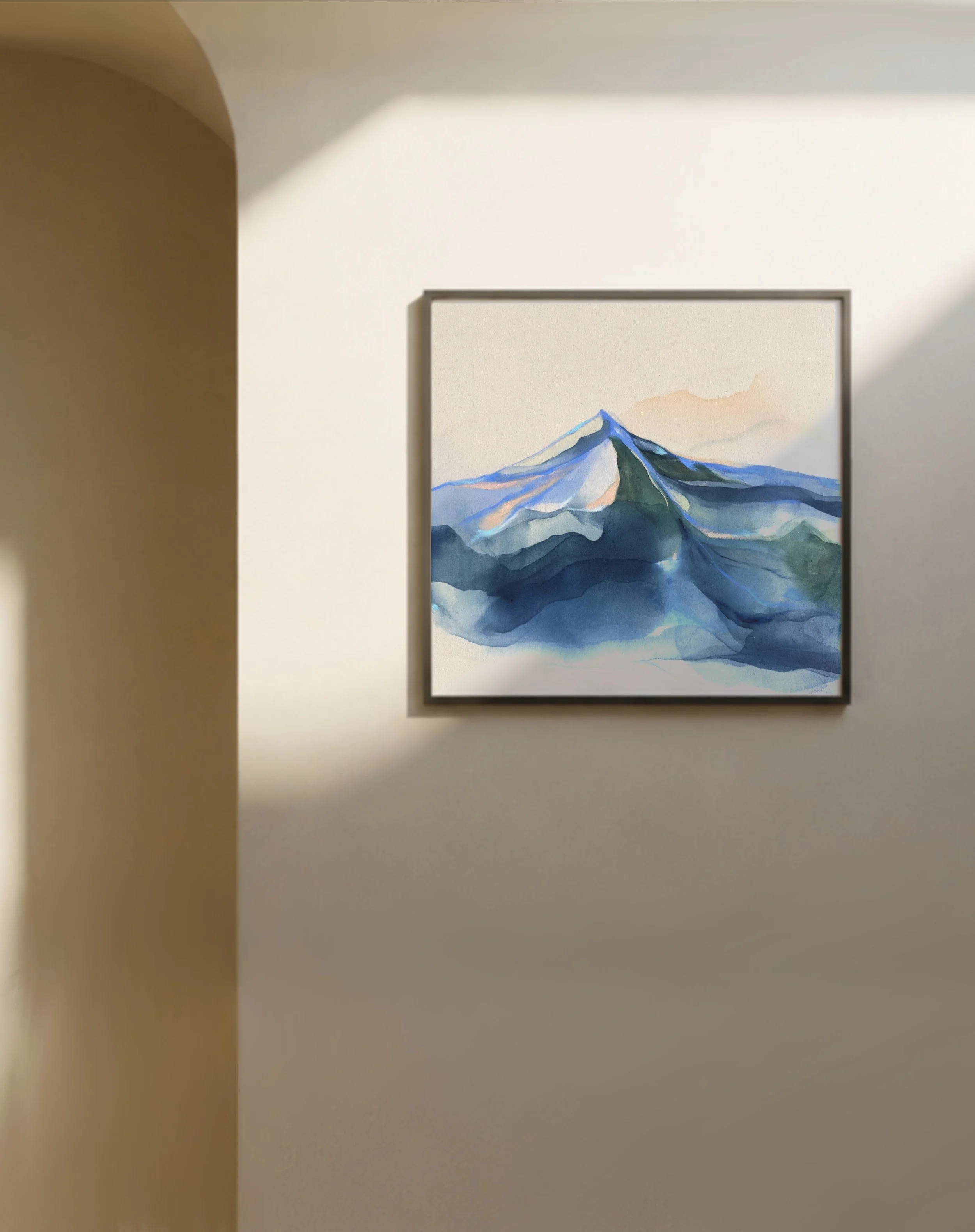 A framed watercolor landscape painting of mountains with green and blue hues, leaning against a light-colored wall on a wooden floor, with sunlight casting shadows on the wall.
