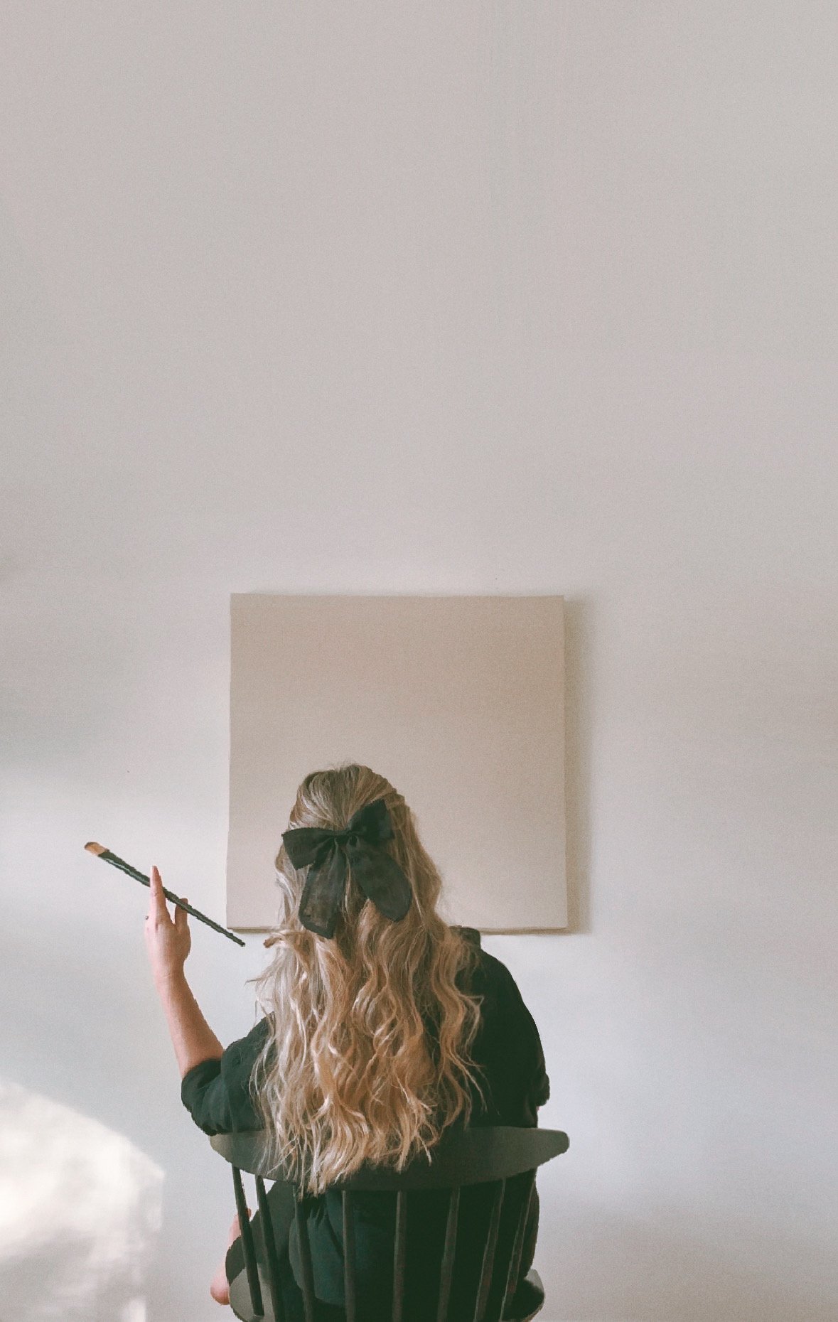 A woman with long wavy hair tied with a large black bow, sitting on a black chair, holding a paintbrush in her right hand, facing a blank canvas on a white wall.
