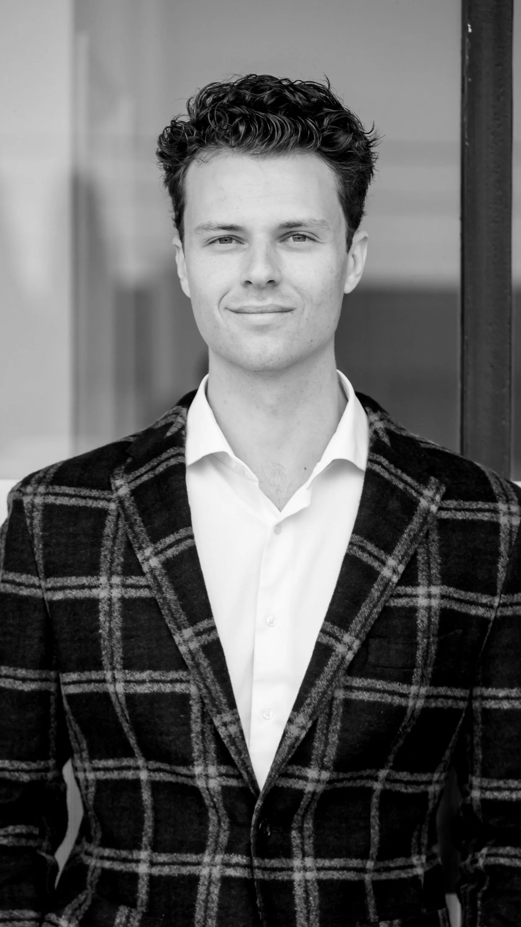 A young man with dark, curly hair dressed in a white collared shirt and a dark plaid blazer, standing outdoors against a blurred background.