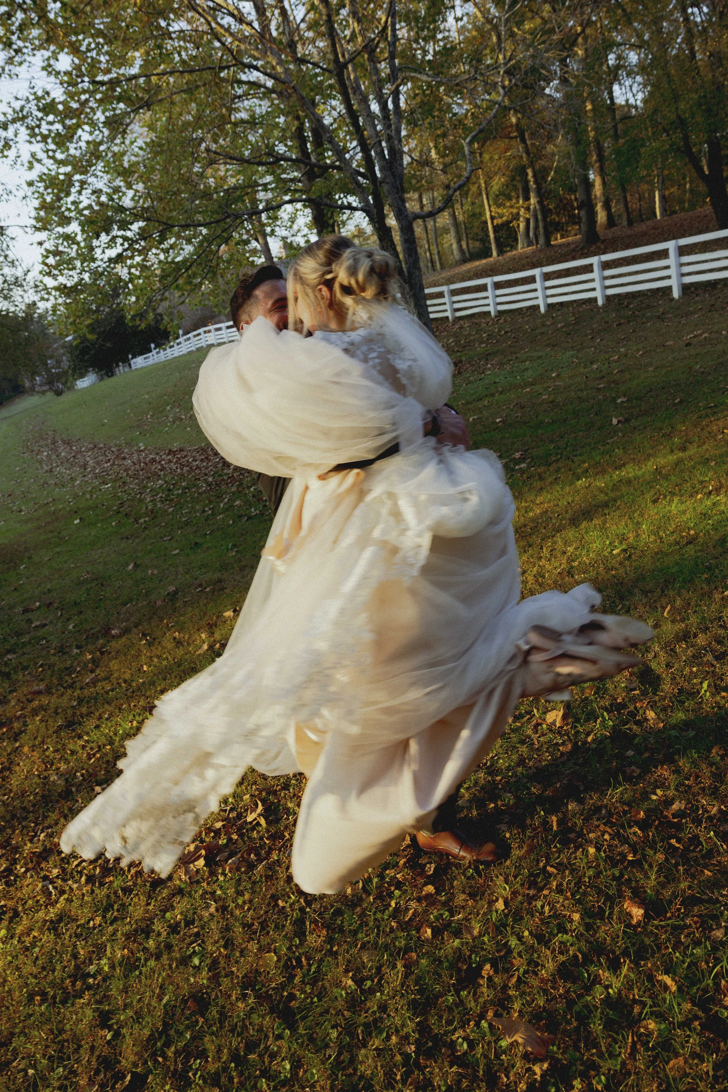 A bride and groom celebrating outdoors, with the groom lifting the bride in a joyful embrace, surrounded by trees and a white fence in the background.