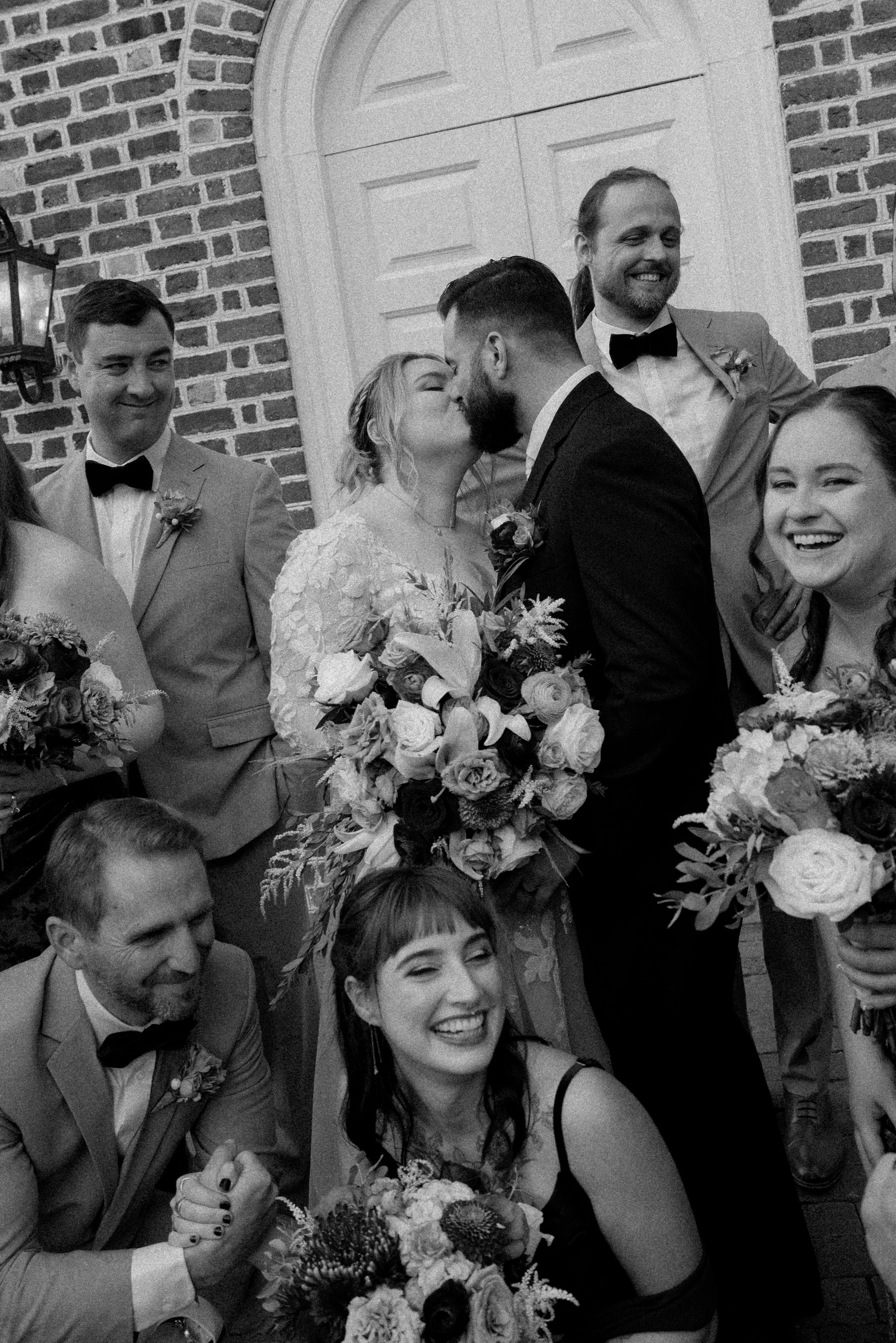 Black and white photo of a wedding group with a bride and groom kissing, surrounded by bridesmaids and groomsmen holding bouquets and smiling, in front of a brick building with a white door.