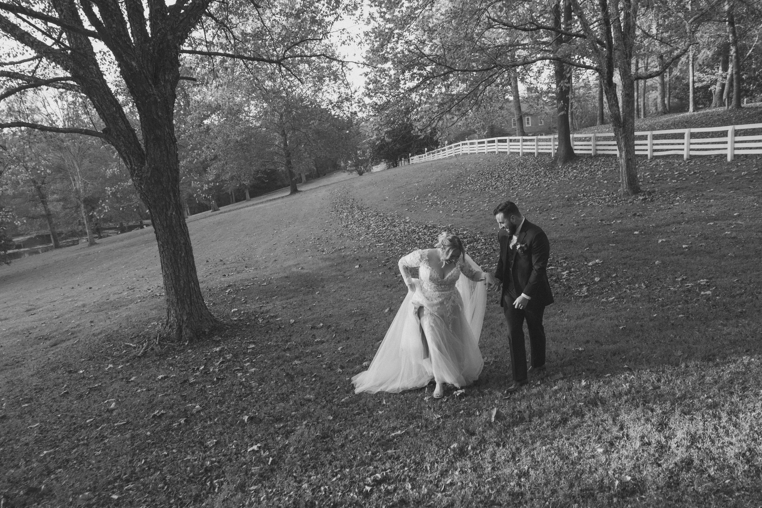 A bride and groom in wedding attire walking on a grassy area in a park with trees and a white fence in the background, black and white photograph.