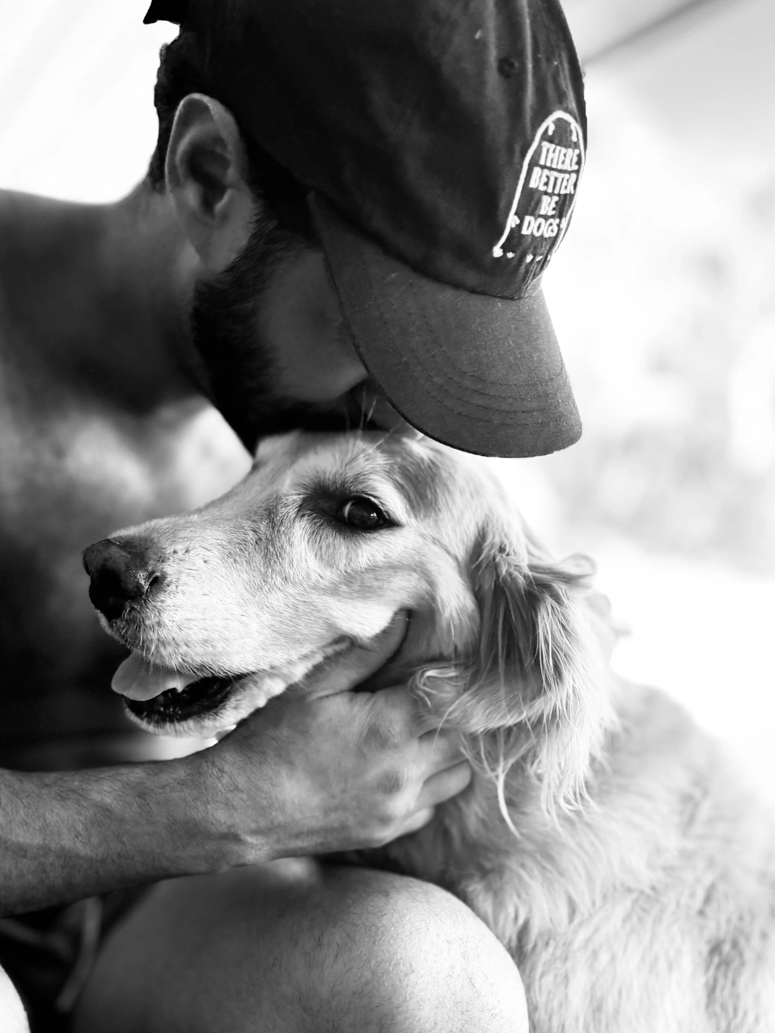 A person wearing a cap with a logo, hugging a happy, light-colored dog with long fur, in black and white.