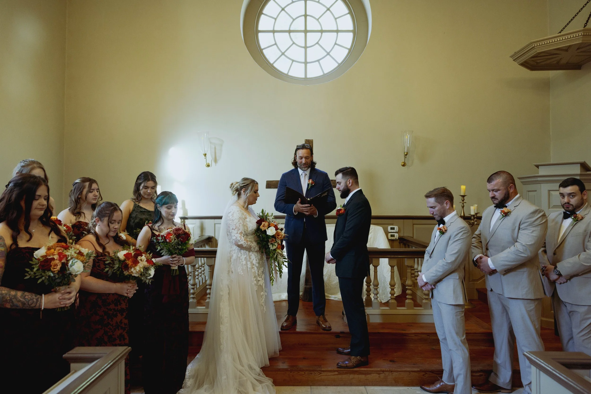 A wedding ceremony with a bride and groom standing before an officiant inside a church. Bridesmaids and groomsmen are standing on either side, holding bouquets and wearing formal attire.