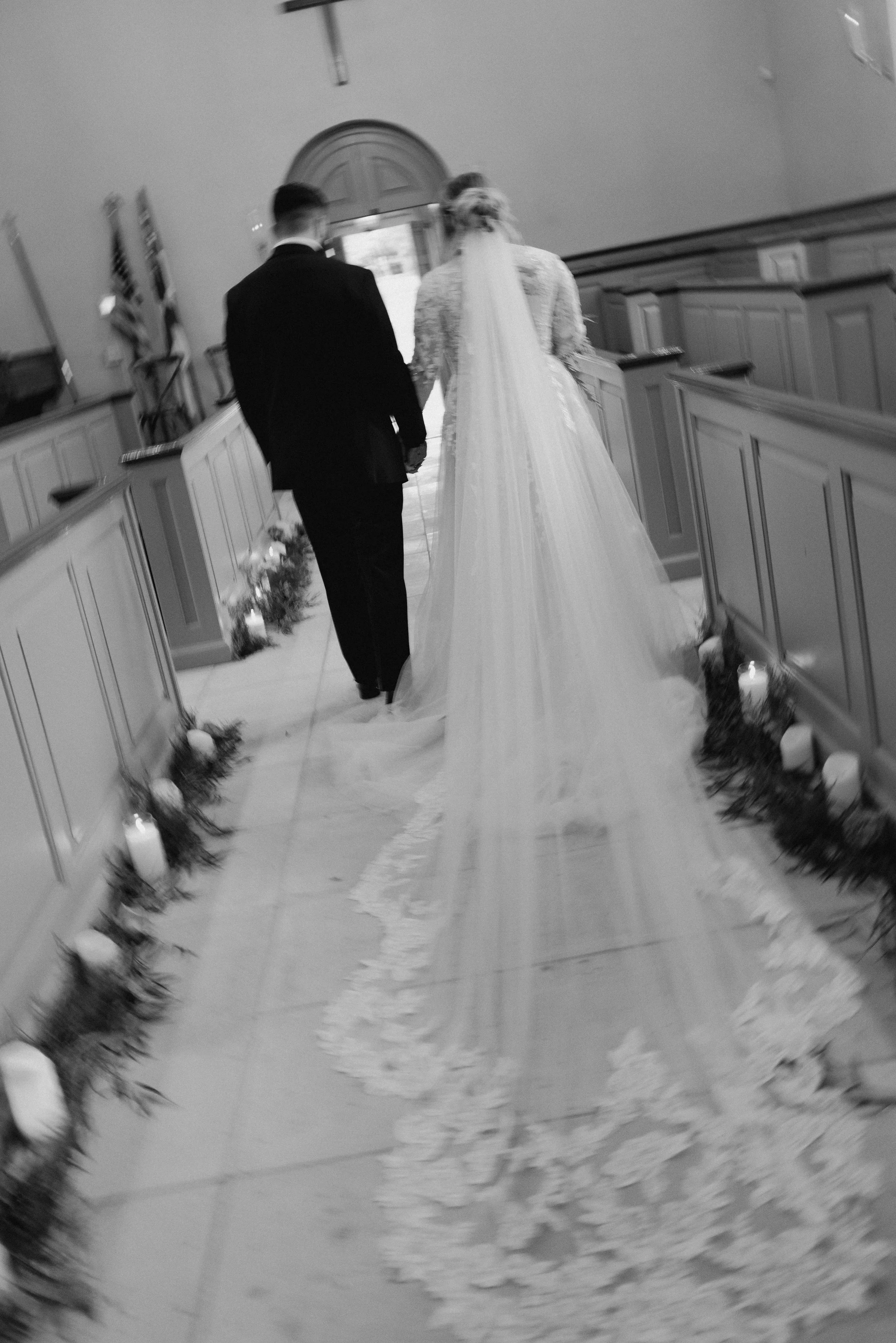 A bride and groom walking down the aisle inside a church or chapel, holding hands, with candles and greenery decorations on the sides.