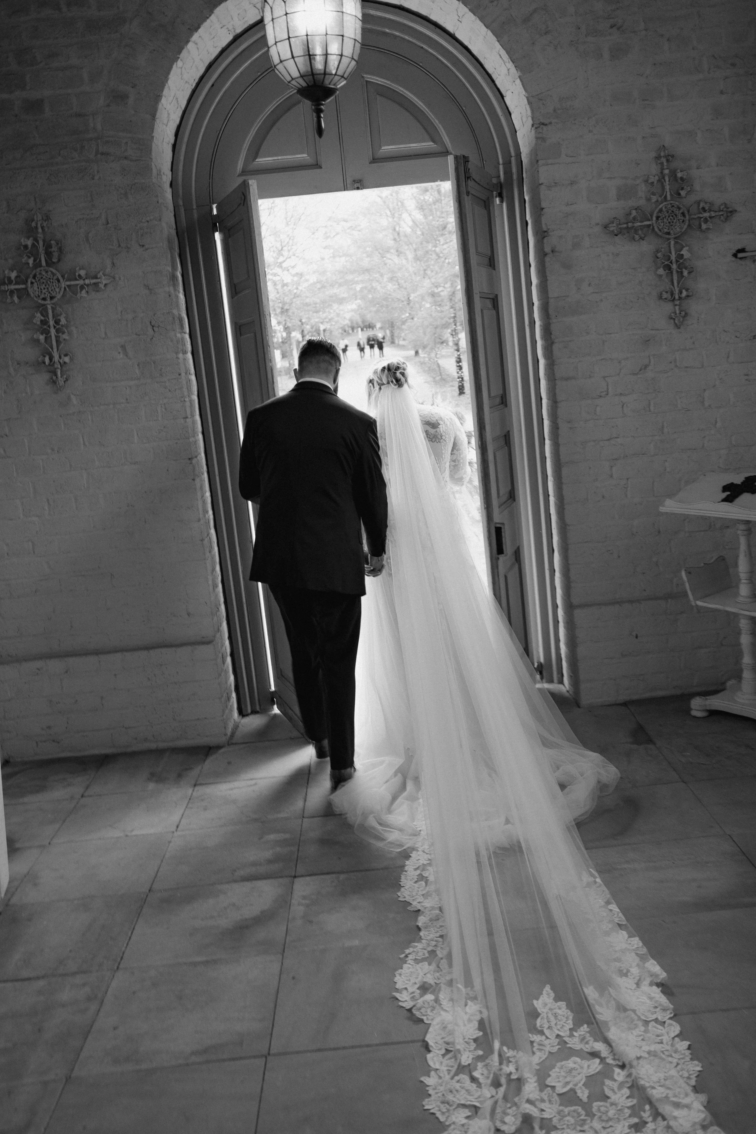 A bride and groom walking out of a church, holding hands, in black and white. The bride is wearing a long, lace-trimmed veil and a wedding dress, while the groom is in a suit, both viewed from behind.
