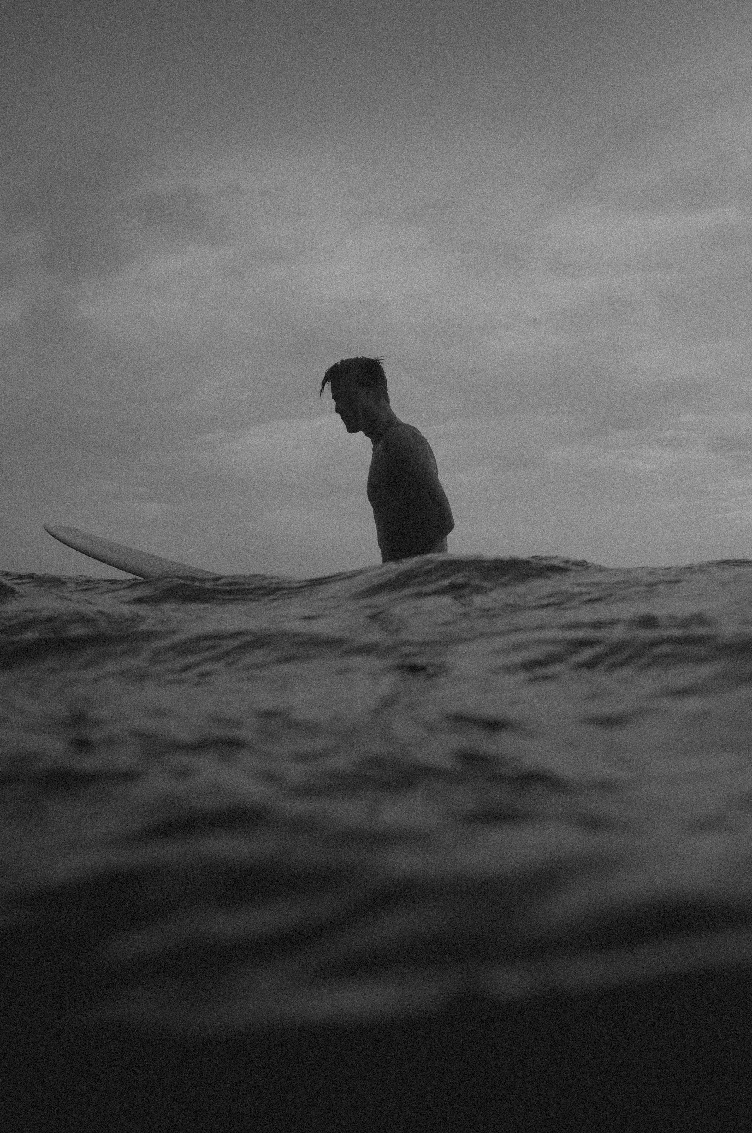 Editorial photo series of a surfer sitting in the water with a surfboard, under a cloudy sky, in black and white.