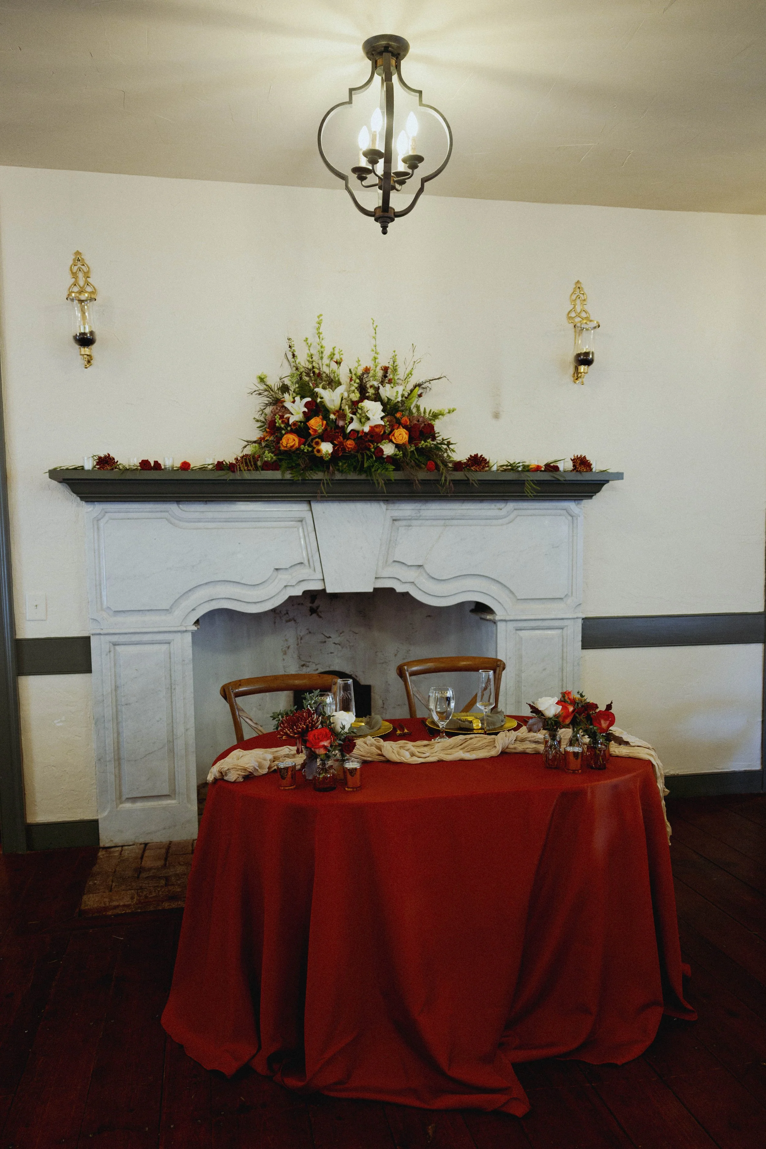 A decorated dining table with a red tablecloth, floral centerpieces, and set glassware in front of a white fireplace with a floral arrangement on the mantel, in a room with a ceiling light and wall sconces.