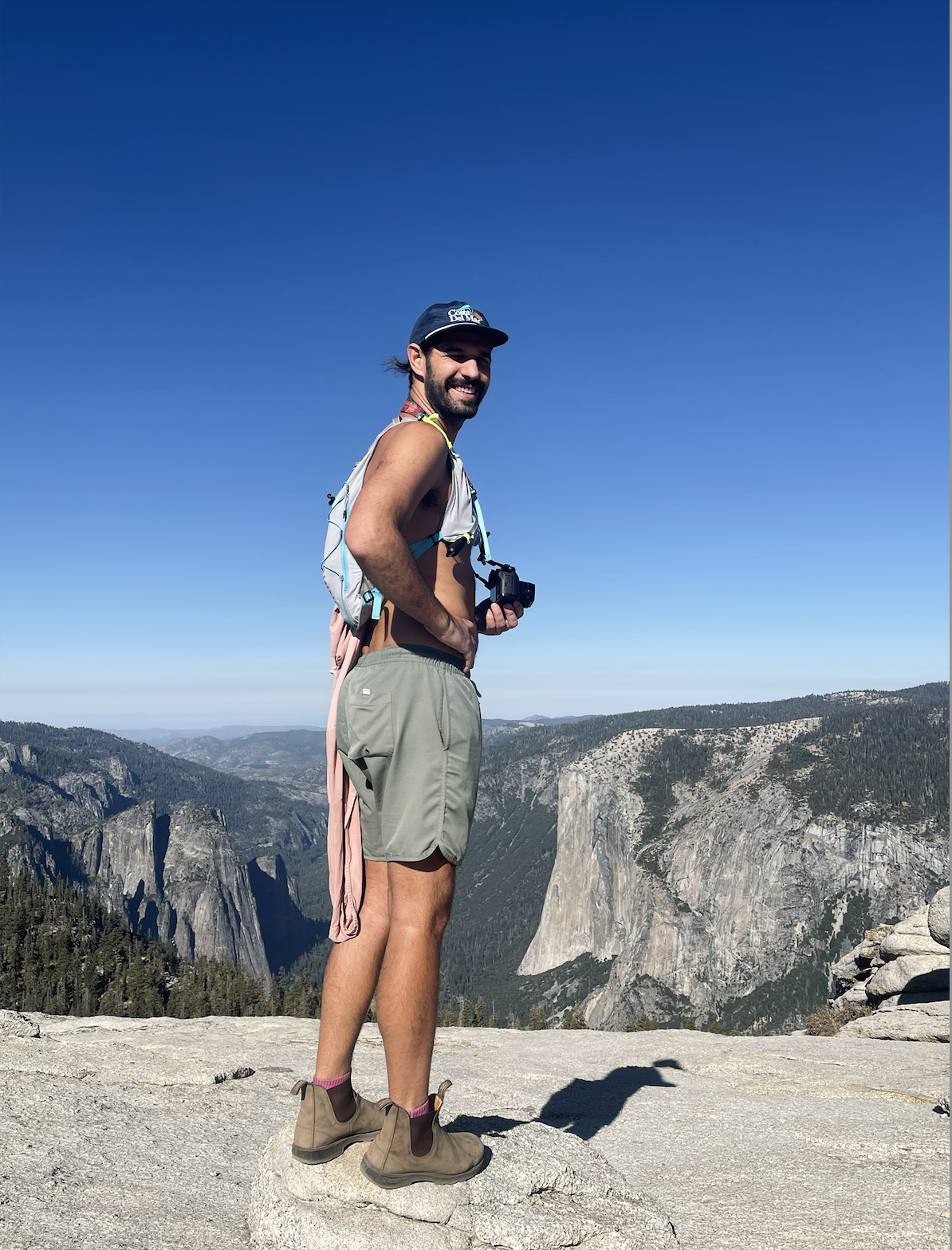 A smiling man standing on a rocky ledge in a mountainous area, wearing hiking shorts, boots, and a hat, with a camera around his neck, overlooking a scenic view of cliffs and valleys.