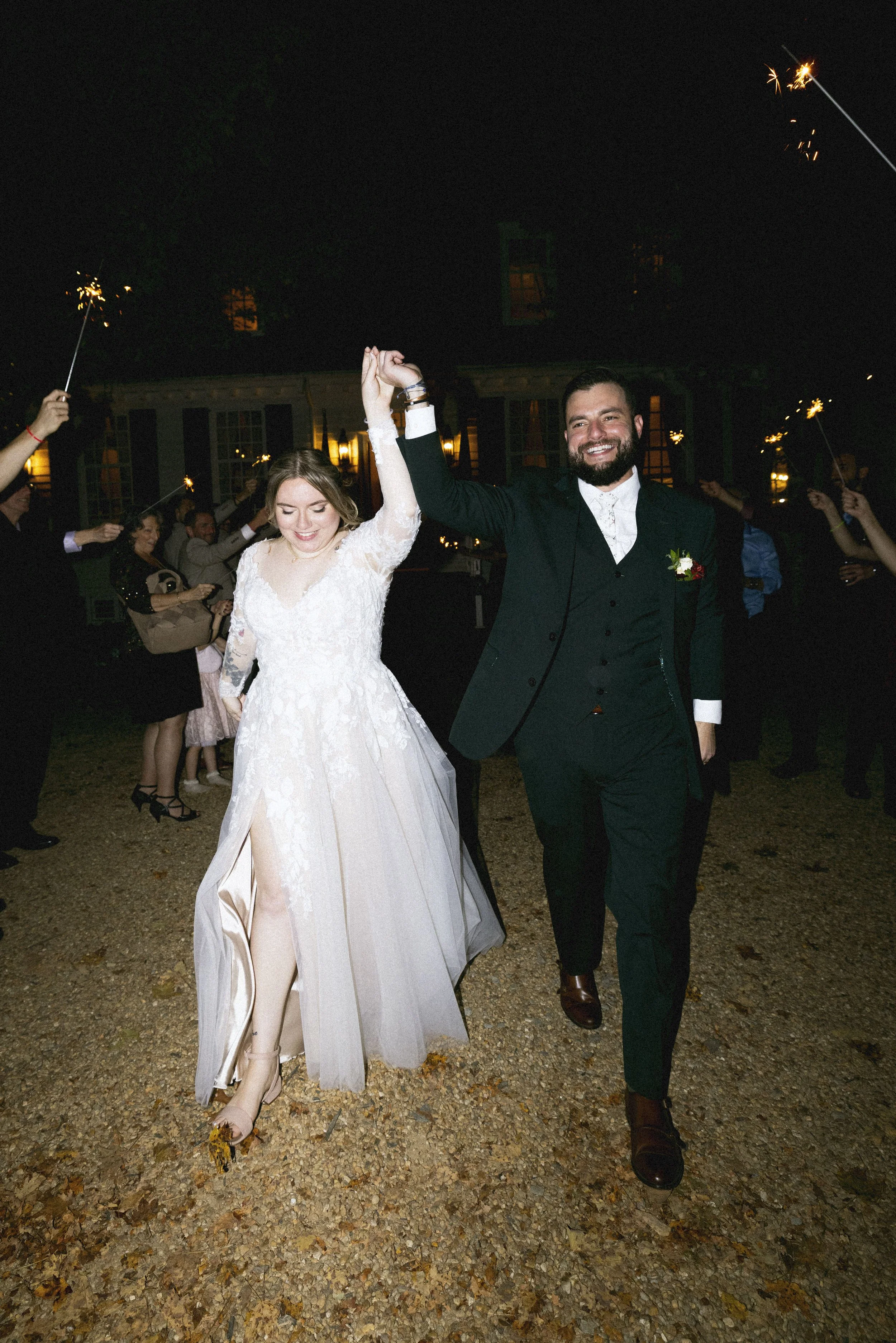A newlywed couple dancing at night wedding reception, the bride wearing a white lace wedding gown and the groom in a dark suit, holding hands and smiling, surrounded by guests holding sparklers outside.