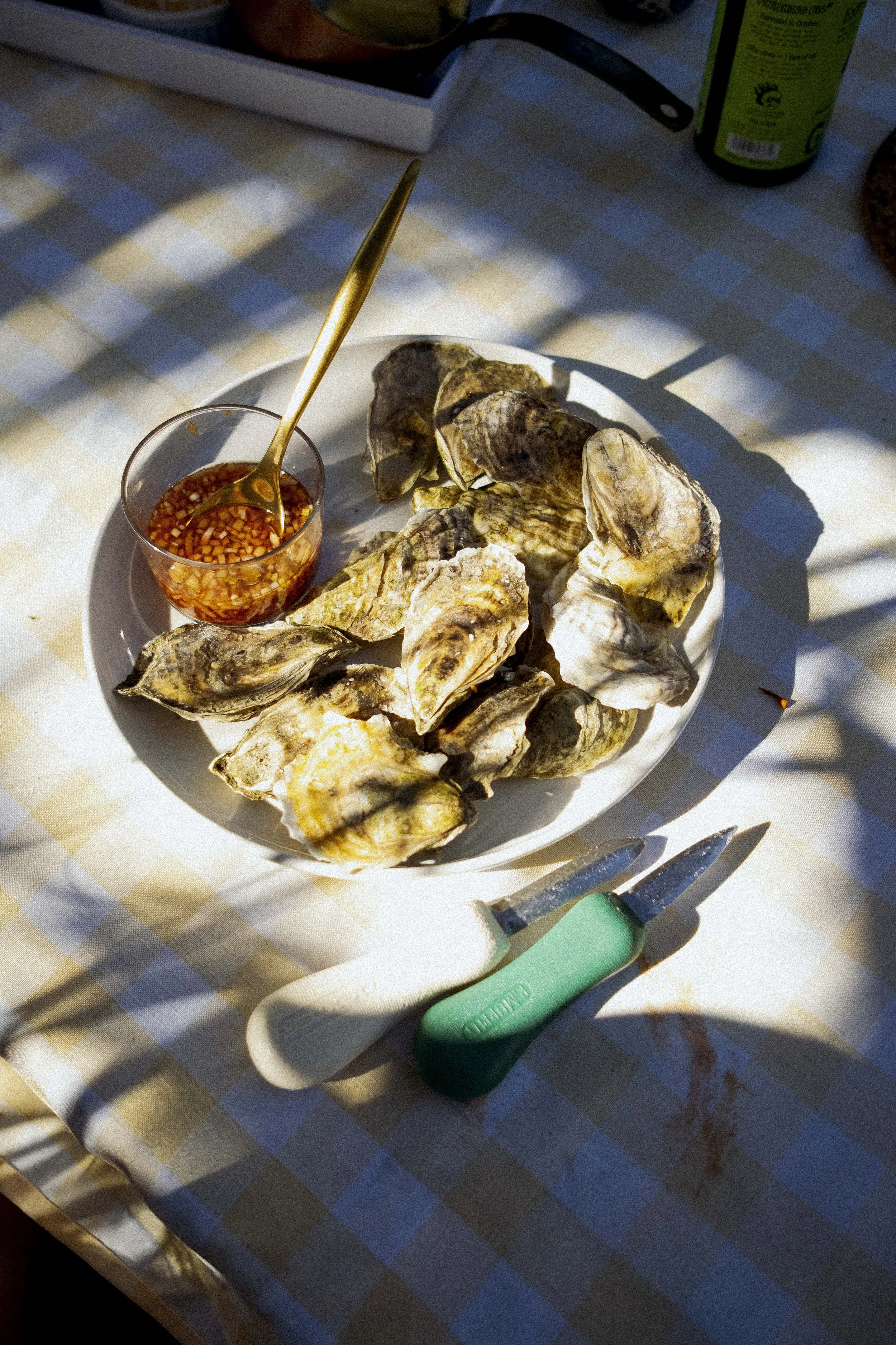 A plate of oysters with a small bowl of cocktail sauce on a checkered tablecloth, with oyster shucking knives nearby.