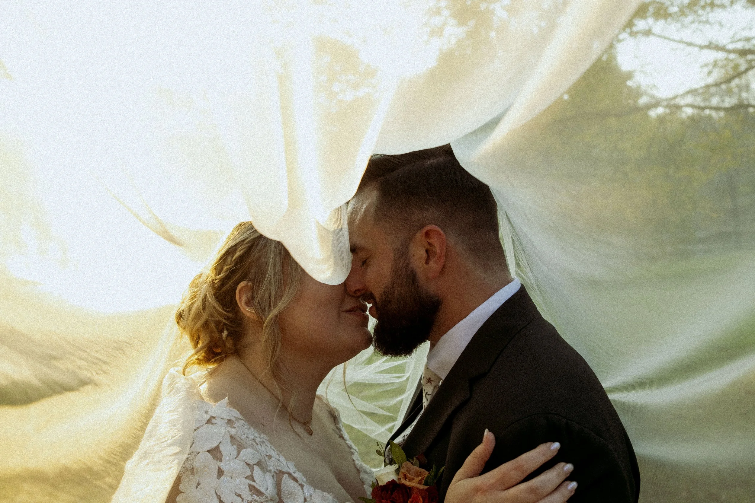 A bride and groom with foreheads touching under white fabric, embracing during their wedding day. 