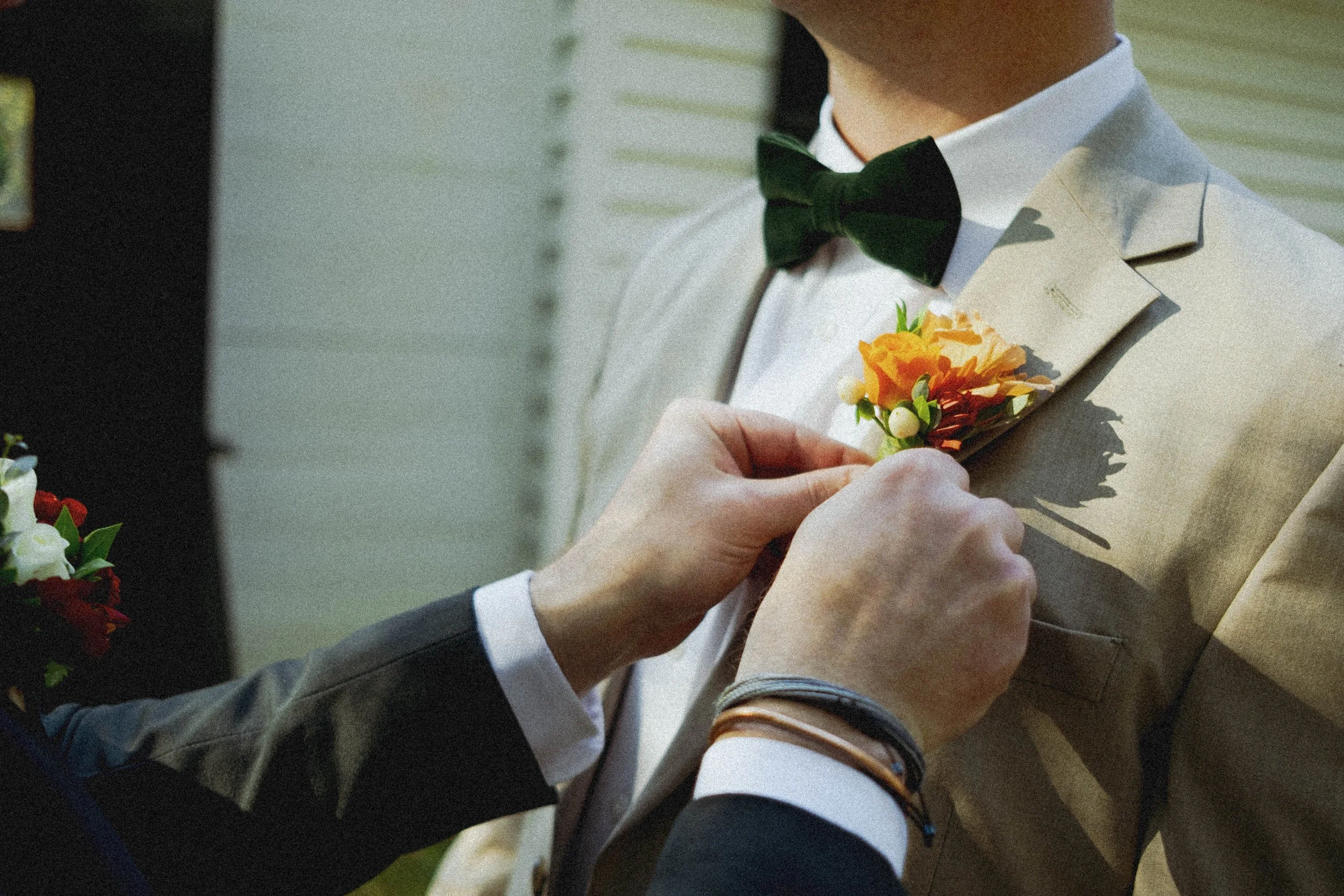 Person in a beige suit with a black bow tie pinning a floral boutonniere onto another person's beige suit.