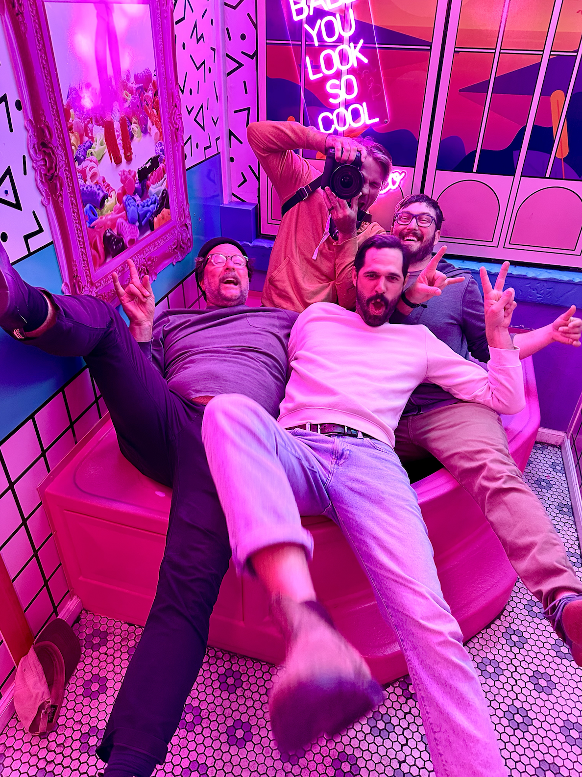 Four men are taking a selfie in a vibrant, neon-lit room with pink and purple lights. They are sitting on a pink bench, making peace signs and expressive faces. The room features colorful wall art, a neon sign that reads 'BAD YOU LOOK SO COOL,' and a mirror showing plush toys.