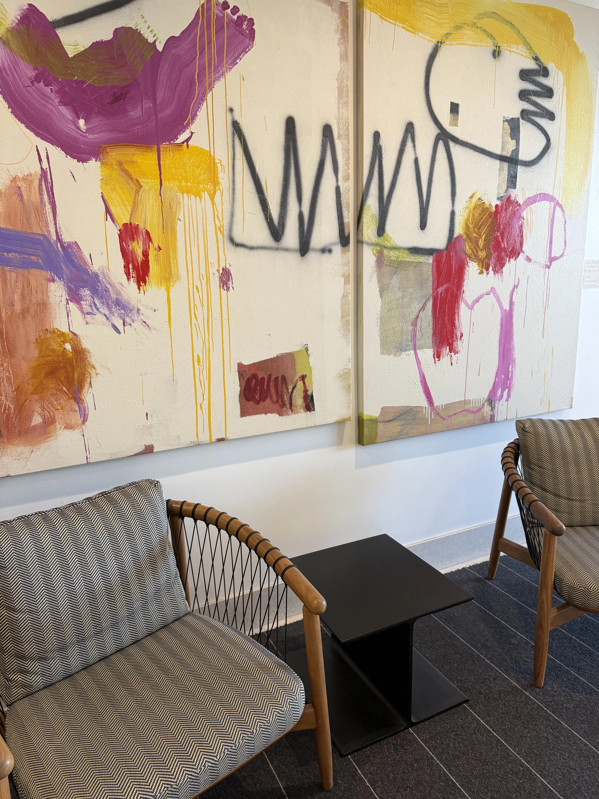 Modern seating area with two upholstered chairs that have wooden armrests and black and white zigzag patterned cushions, a small black table, and large abstract artwork on the wall featuring colorful paint splashes and graffiti-style text.