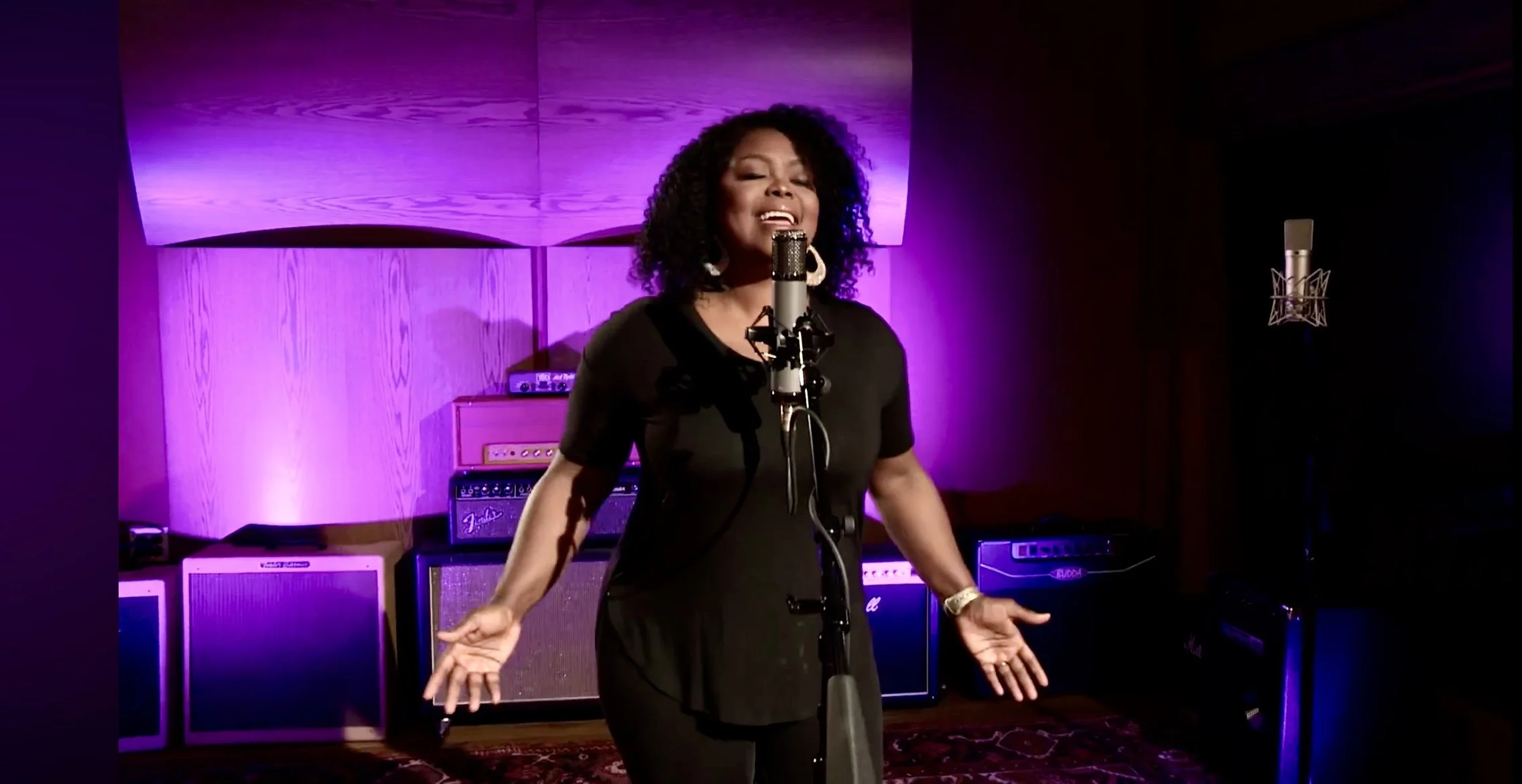 A woman singing into a microphone in a recording studio with purple lighting and guitar amplifiers in the background.