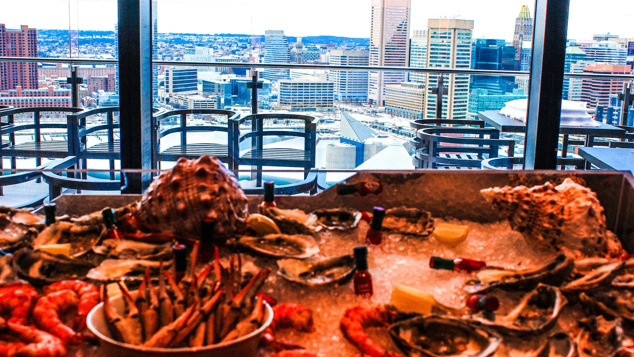 Seafood platter with oysters, lobster, shrimp, and crab claws on a table overlooking a city skyline with tall buildings and snow on rooftops.