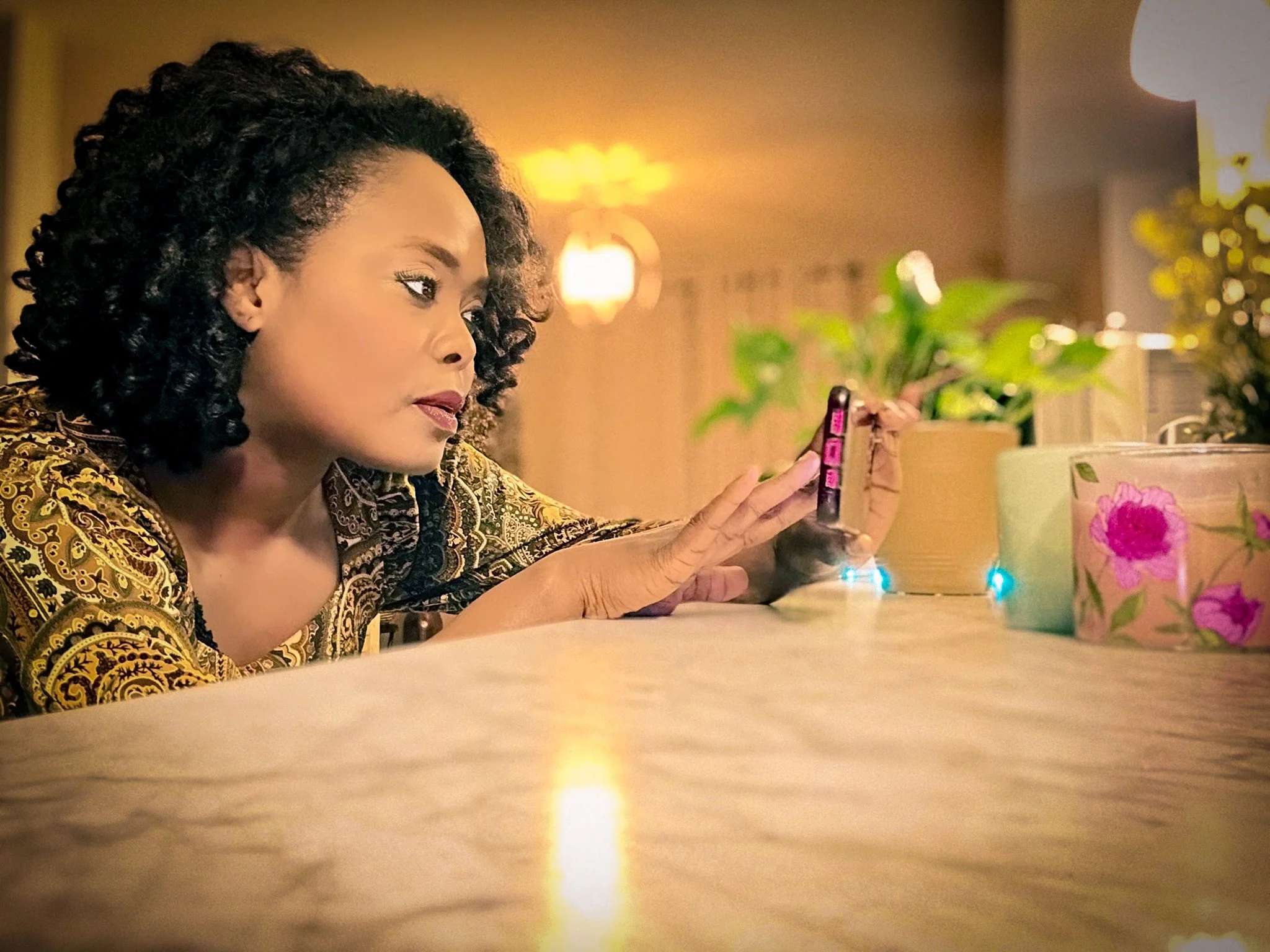 Woman with curly hair in a patterned shirt lying on a table, looking at her phone, with potted plants and a lit lamp in the background.