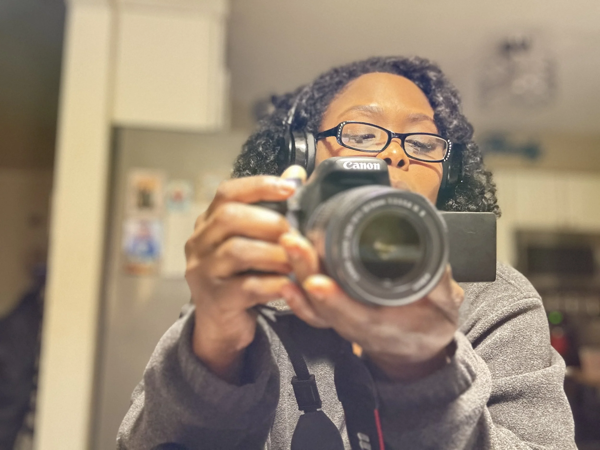 A woman with curly hair, glasses, and earphones taking a selfie with a Canon camera in a kitchen.